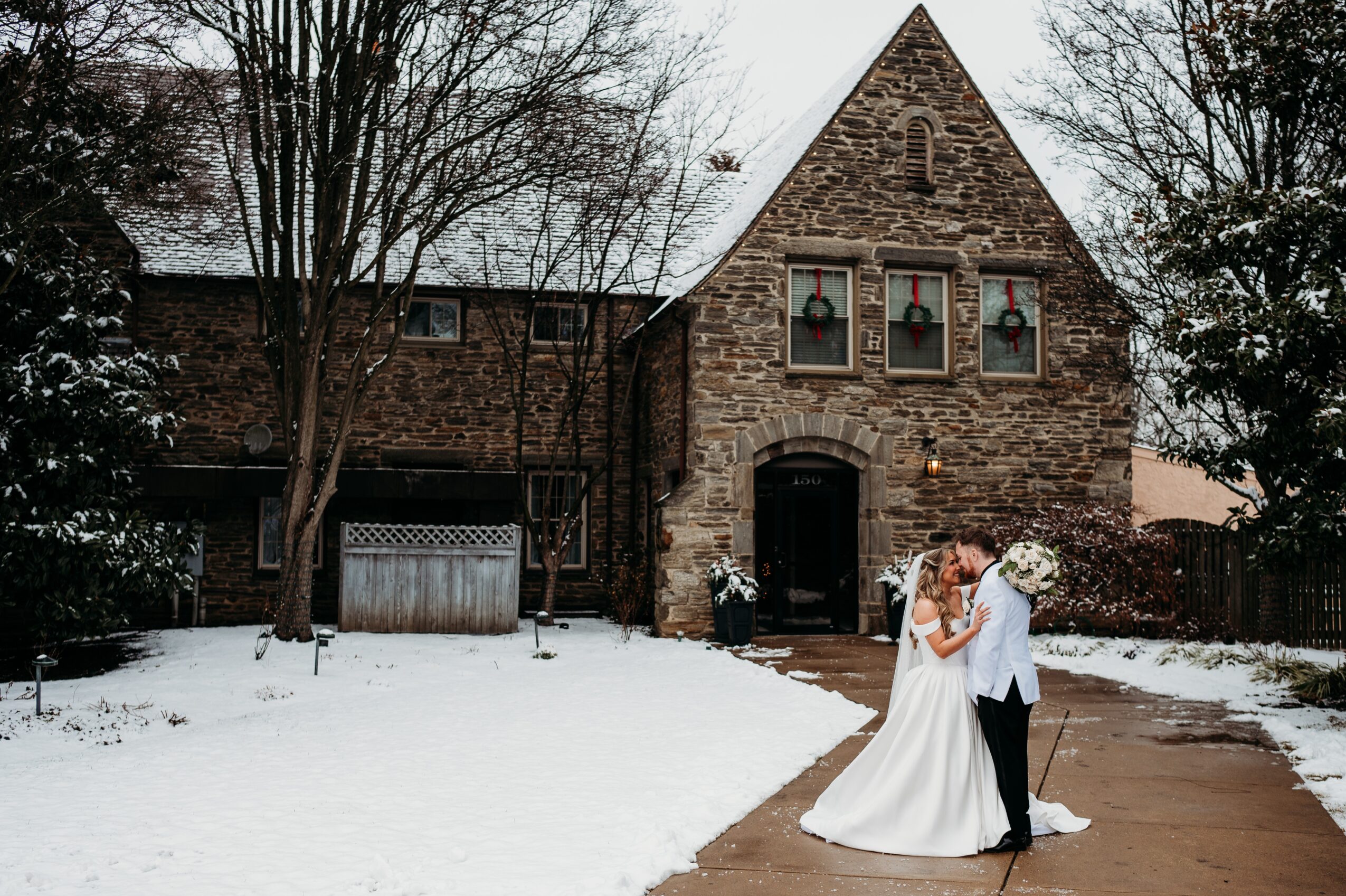 A bride and groom kiss outside a stone house with Christmas decorations in the windows, snow on the ground.