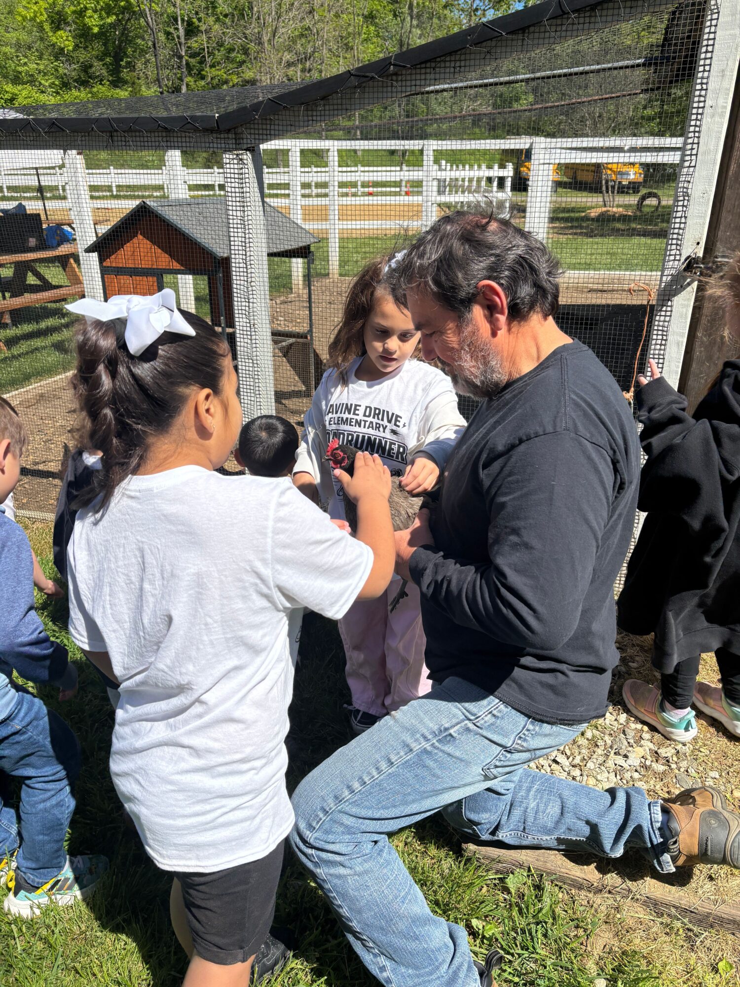 Two people kneeling on grass, talking outdoors near a fence and trees, with a person in a white shirt and a person in a black jacket.