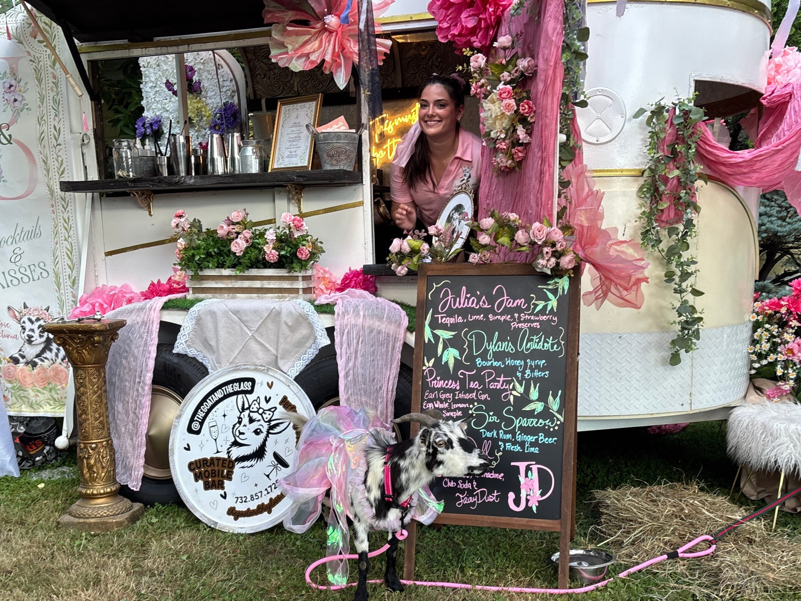 Woman smiling at a decorated outdoor event with pink flowers, a chalkboard menu, and a white caravan, surrounded by pink and white decorations.
