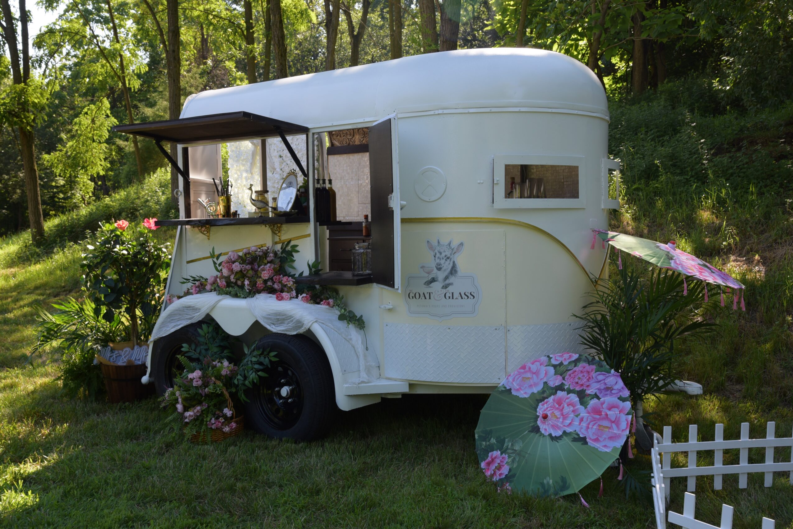 White vintage trailer decorated with flowers and umbrellas, surrounded by greenery and a small white fence.
