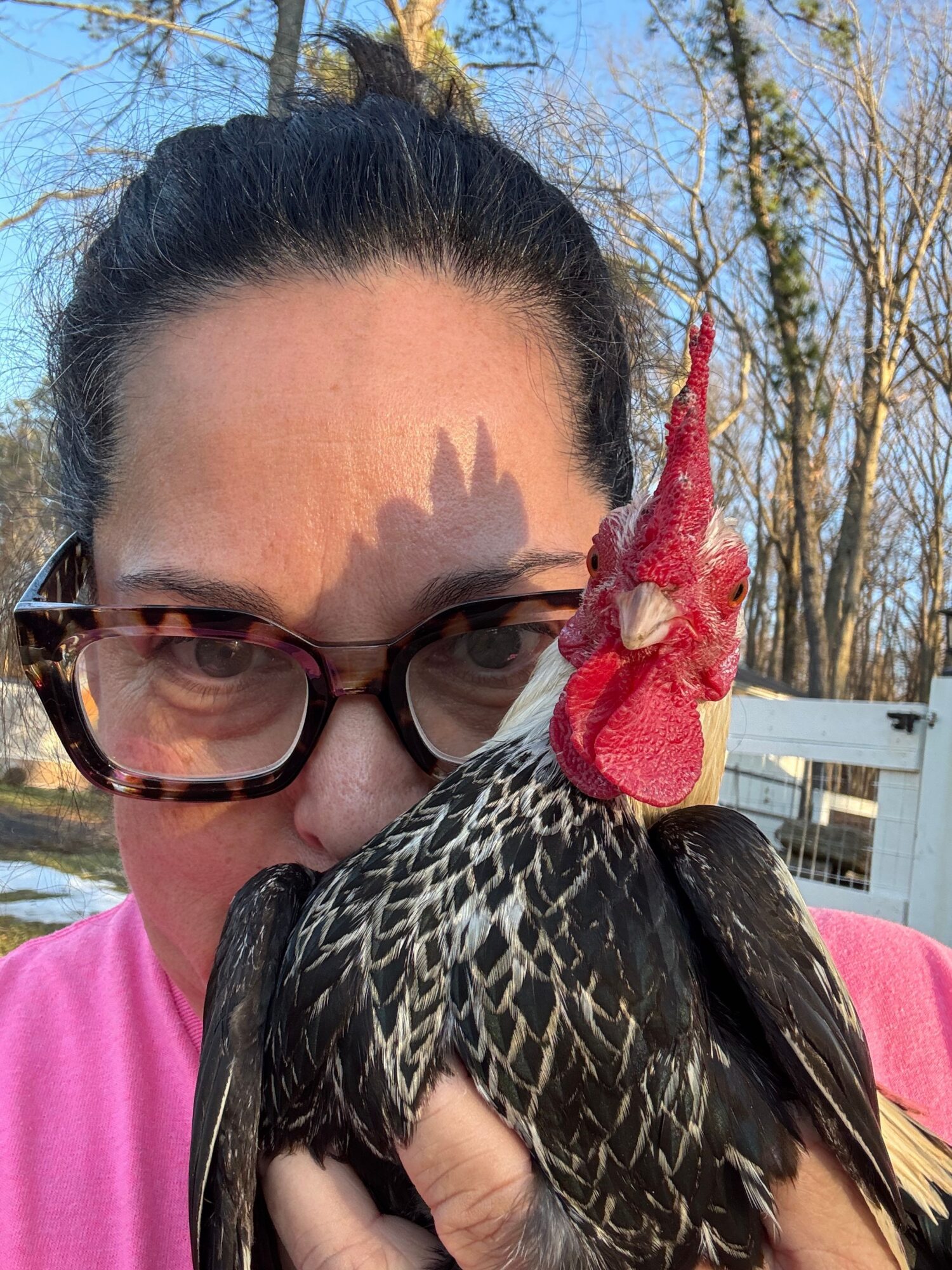 Person with glasses holding a bird close to their face outdoors, with trees and a building in the background.
