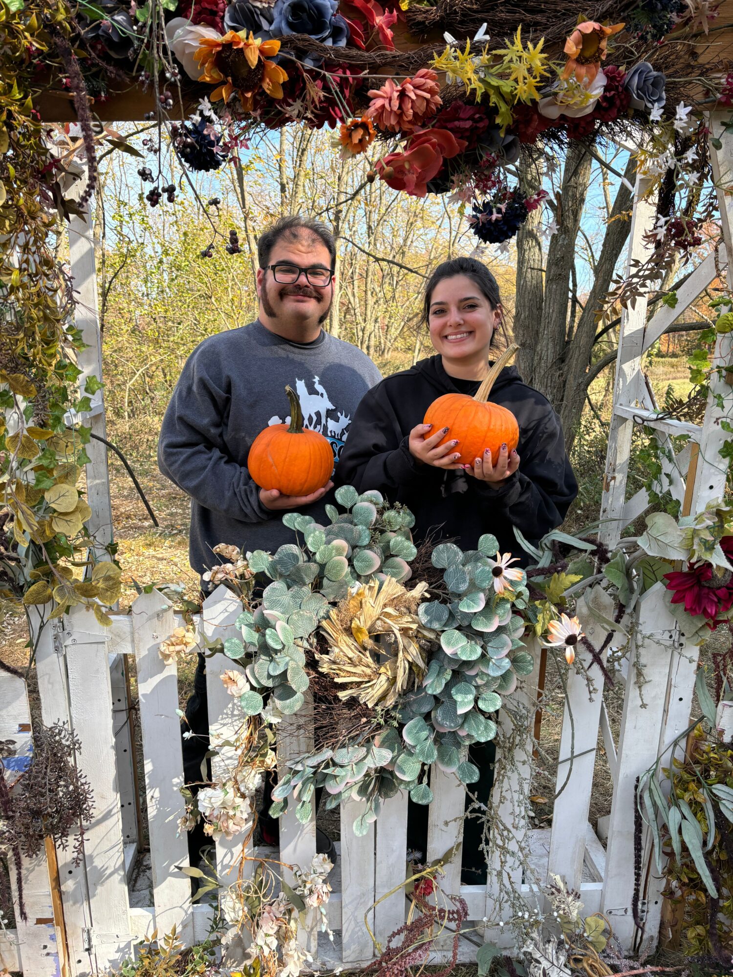 Two people holding small pumpkins in front of a decorated outdoor setting with flowers and plants.