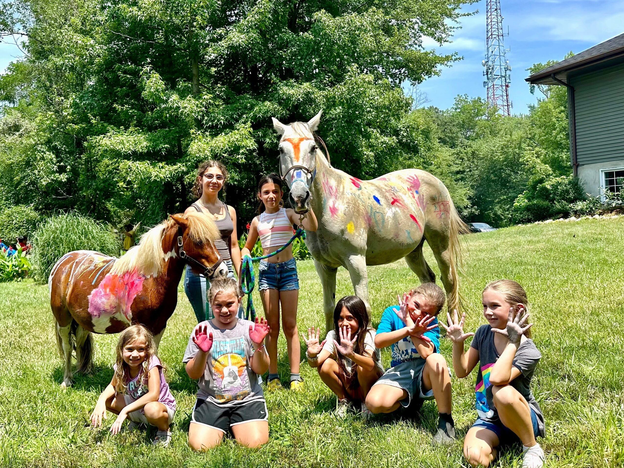 Group of children and two painted horses outdoors on grass with trees and a house in background.