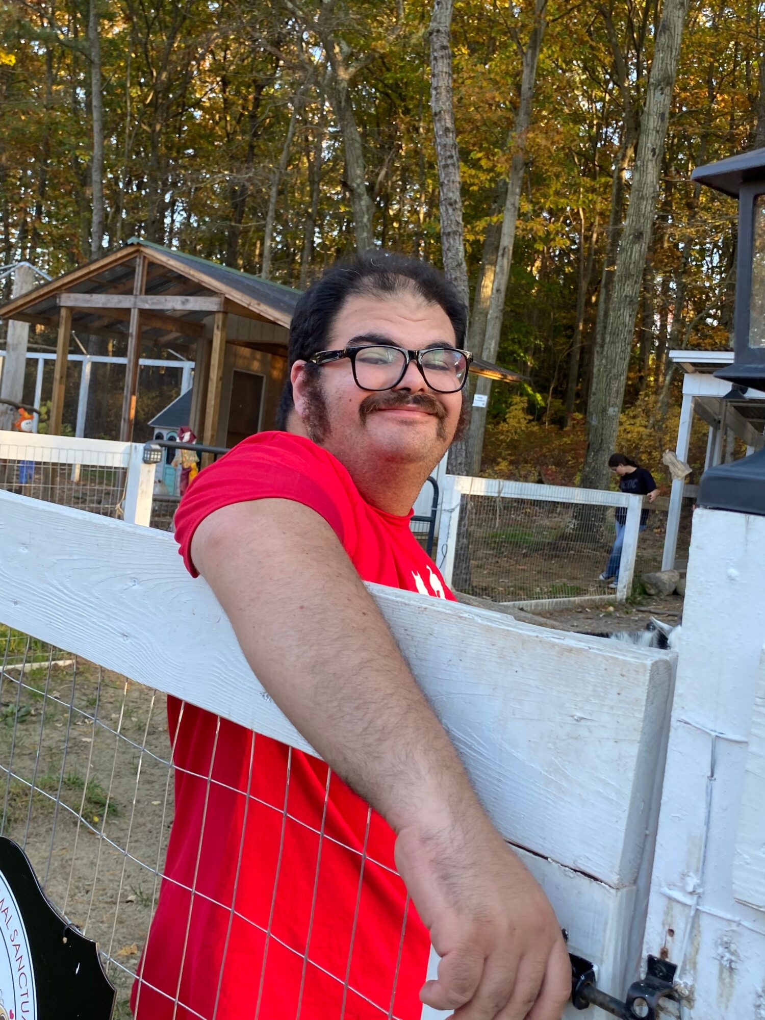 Person with glasses smiling, sitting at a white table outdoors, wearing a red shirt, with trees in the background.