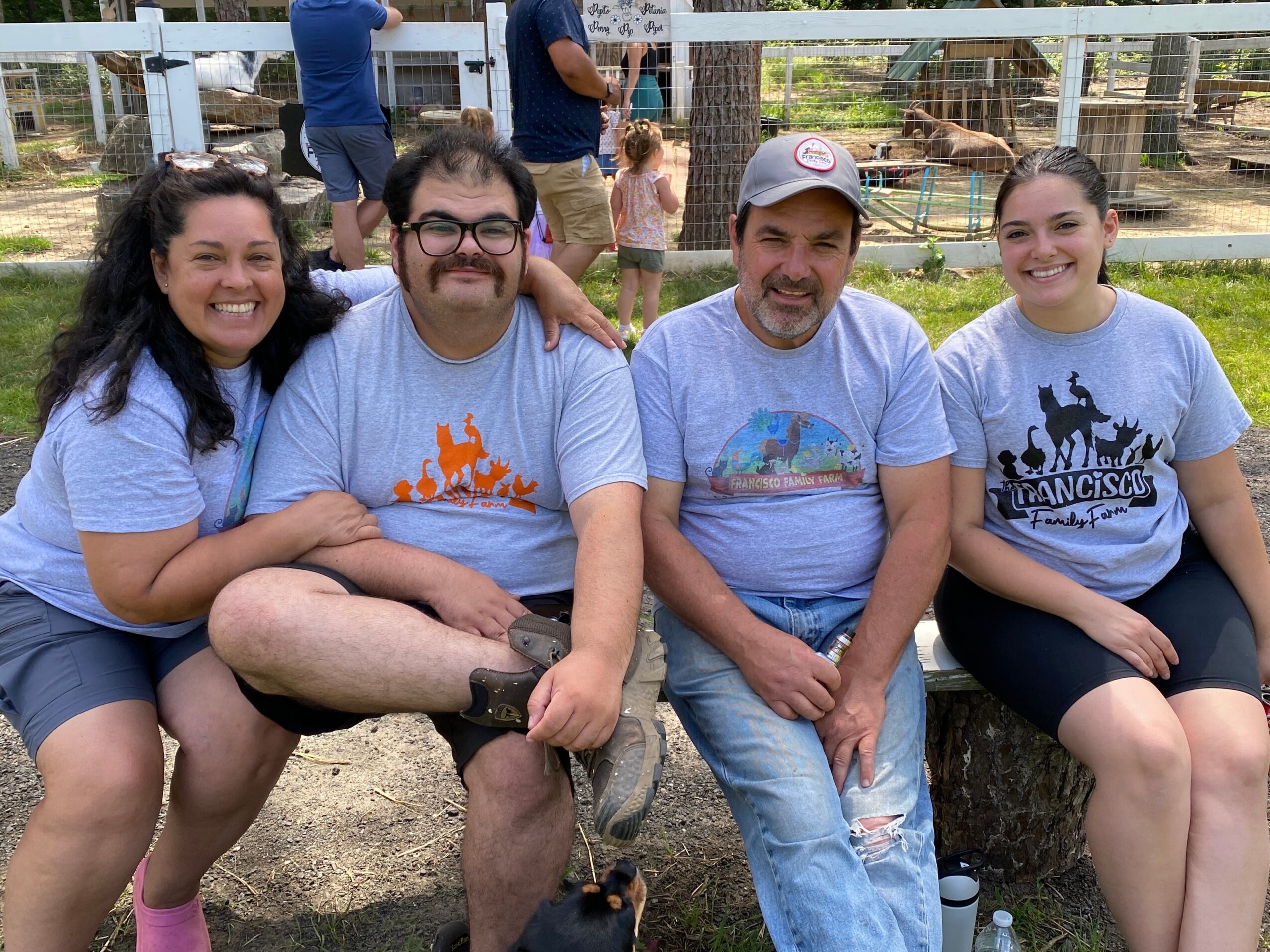 Four people sitting outdoors, smiling, with a fence and animals in the background, wearing casual clothes and T-shirts.