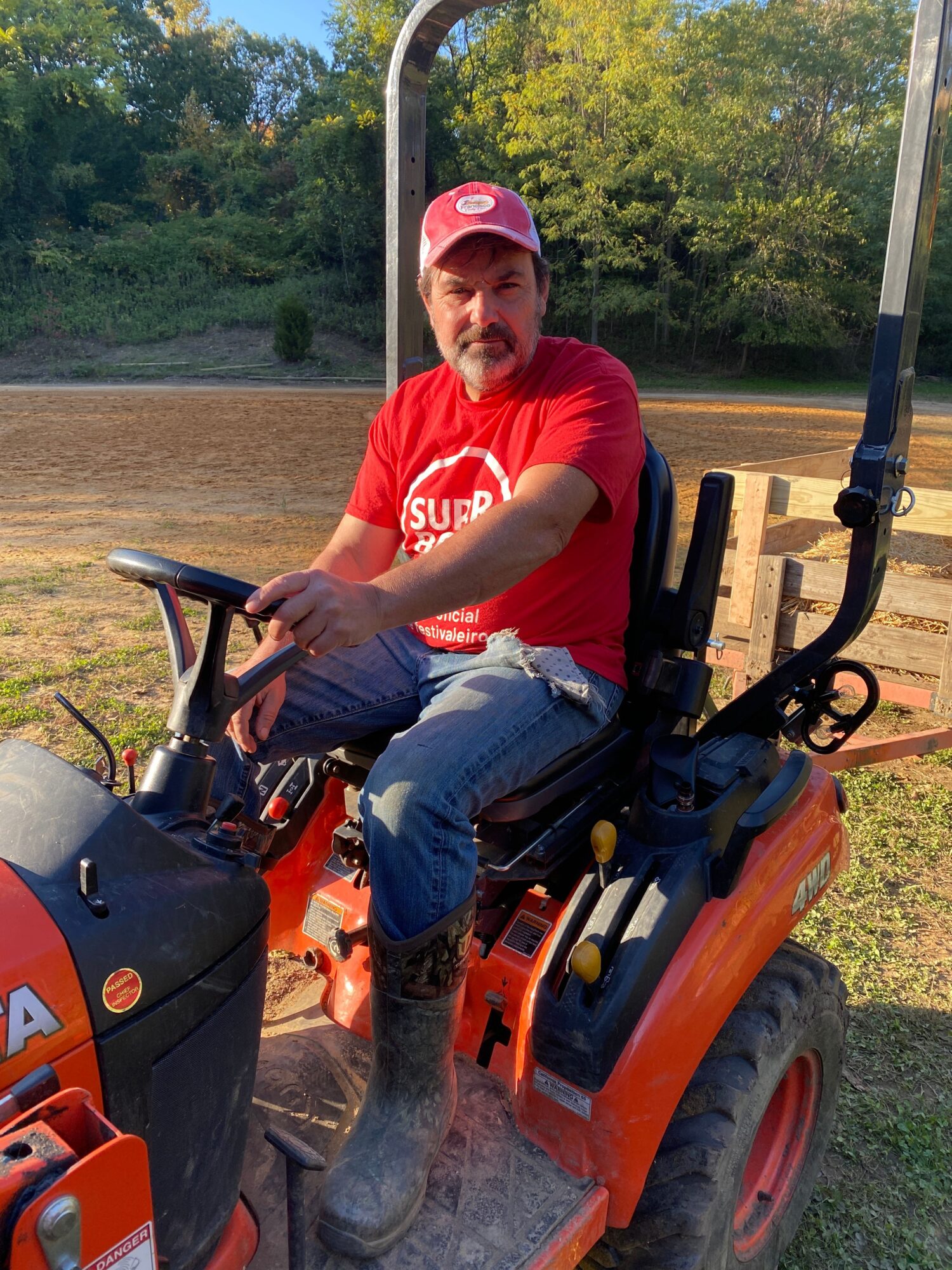 Person wearing a red shirt and pink cap sitting on an orange tractor outdoors, with trees and grass in the background.