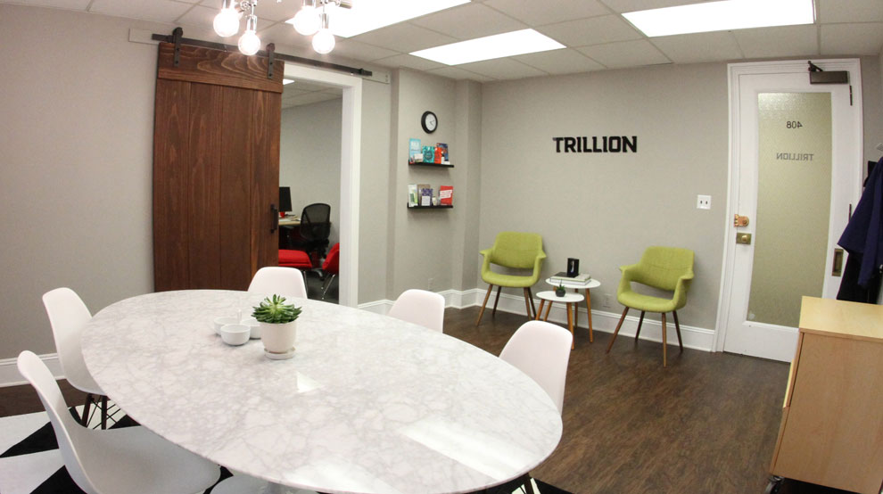 Office meeting room with a round table, chairs, and a wall with shelves and chairs, with a door labeled 'TRILLION'.