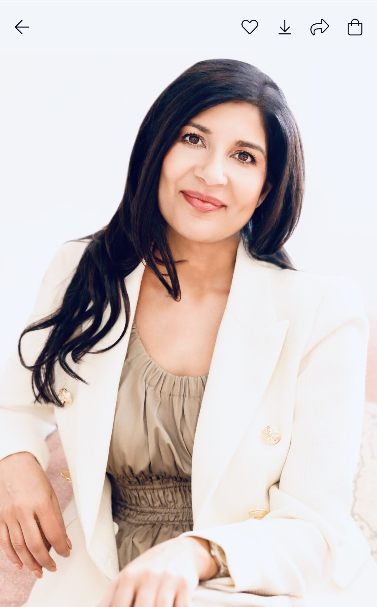 Woman with long dark hair wearing a beige top and white blazer, smiling, against a light background.