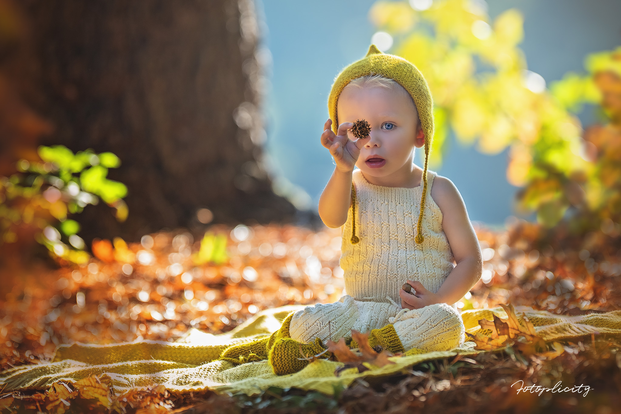 Child sitting outdoors on a blanket, wearing a yellow hat and sweater, with autumn leaves around.