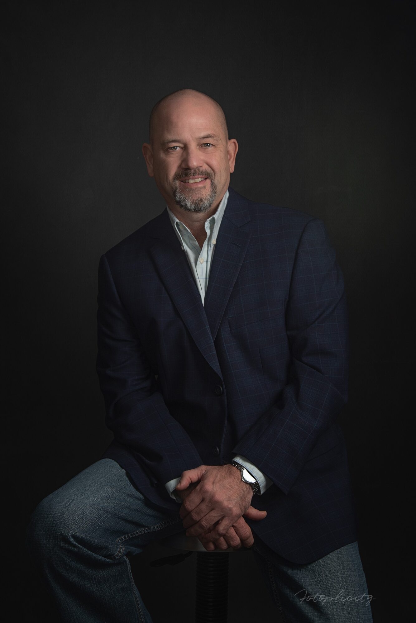 Man with a beard and bald head wearing a dark blazer and white shirt, sitting against a dark background.