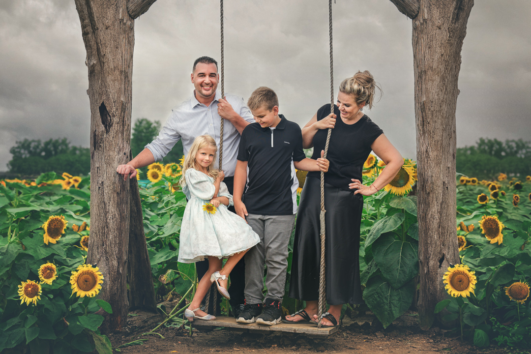 Family of four standing on a swing between two trees in a sunflower field, smiling and posing for the photo.