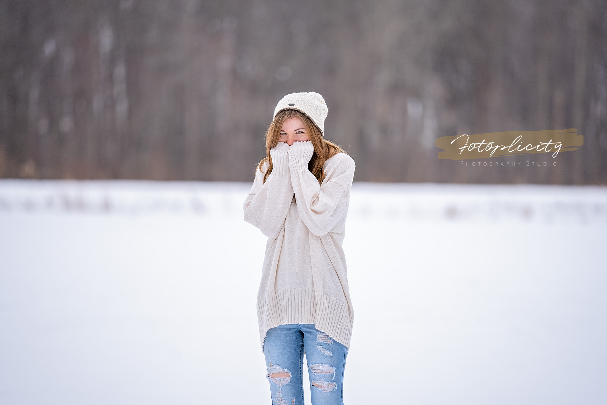 A girl in a white sweater and hat covers her face with her hands outdoors in a snowy landscape.