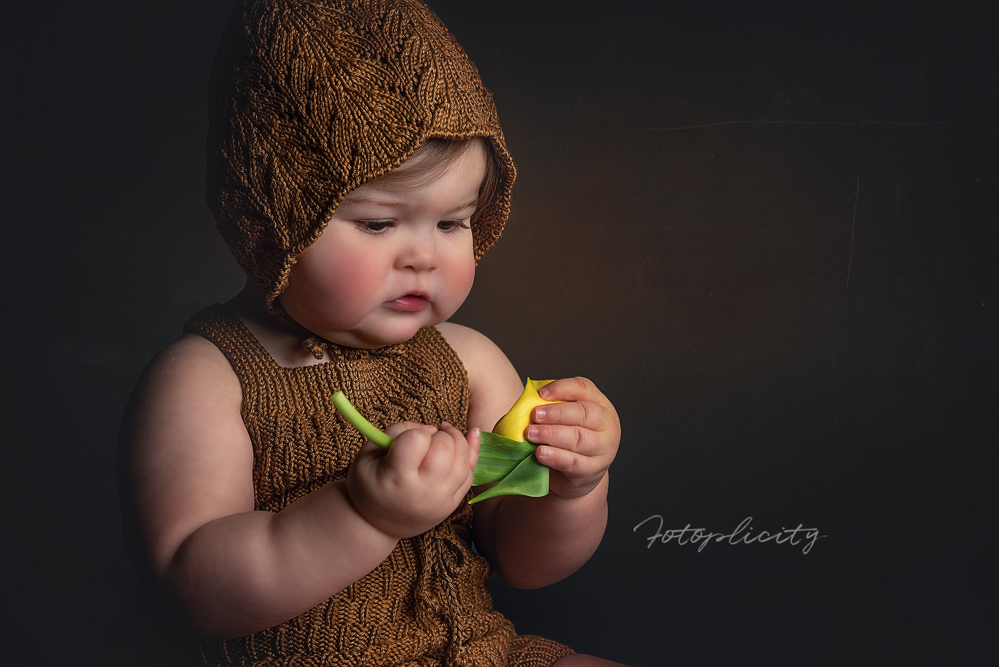 Baby dressed in a brown knitted outfit and hat, holding a yellow and green toy, against a dark background.