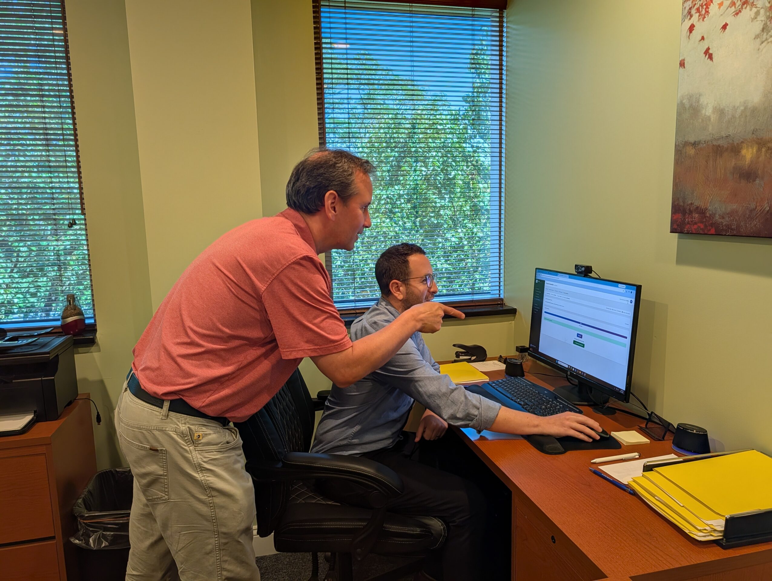 Two men in an office, one standing and pointing at a computer screen, the other sitting at a desk using the computer.