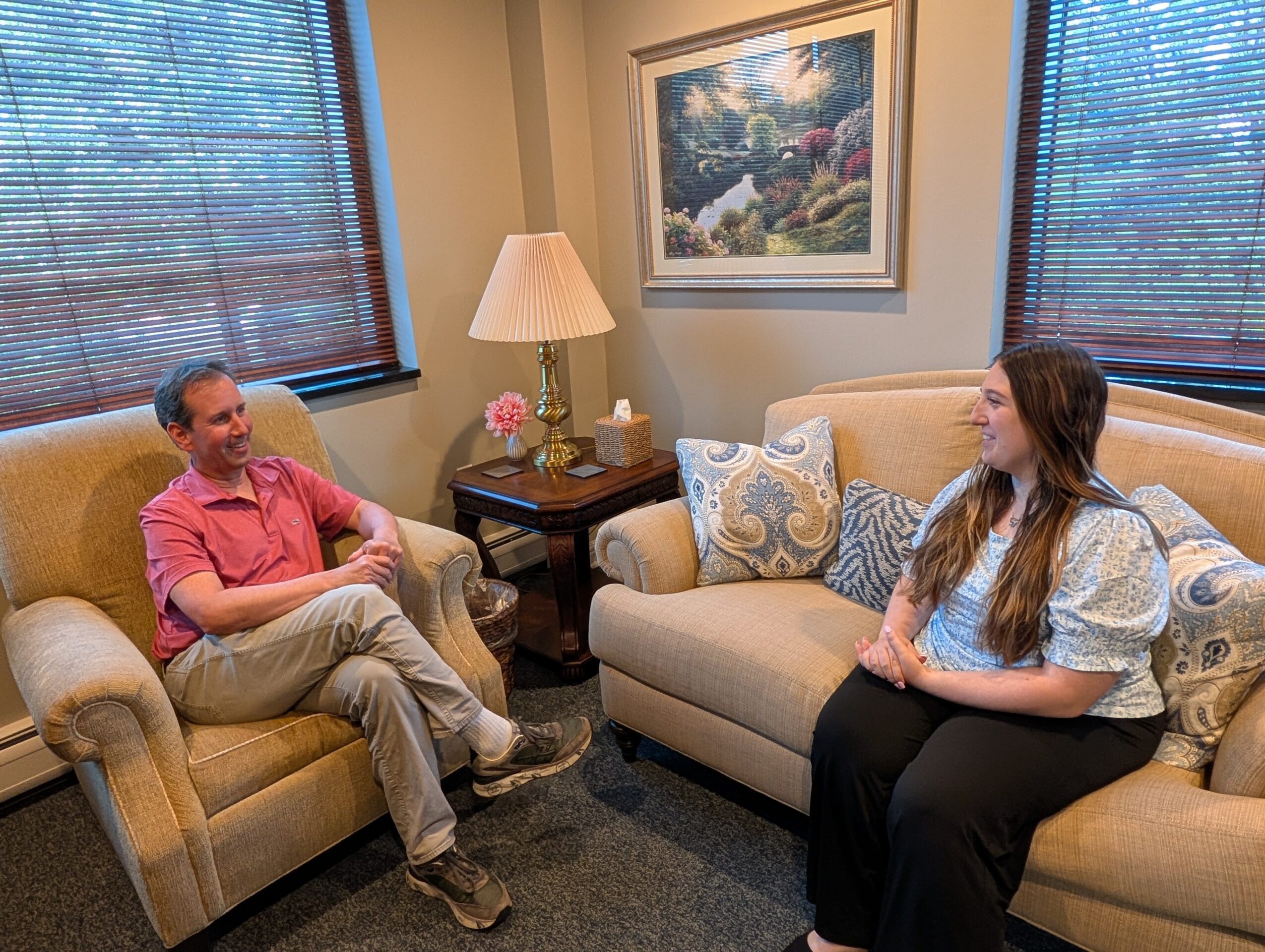 Two people sitting on sofas in a living room, engaging in conversation, with a painting and window blinds behind them.