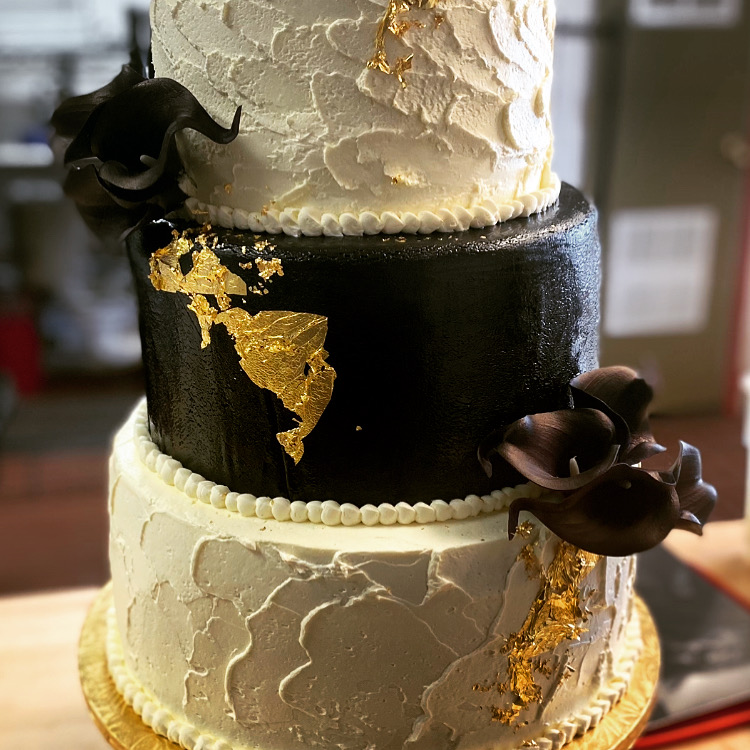 Three-tiered cake with black, white, and gold decorations, featuring gold leaf accents and chocolate flowers, on a gold base.