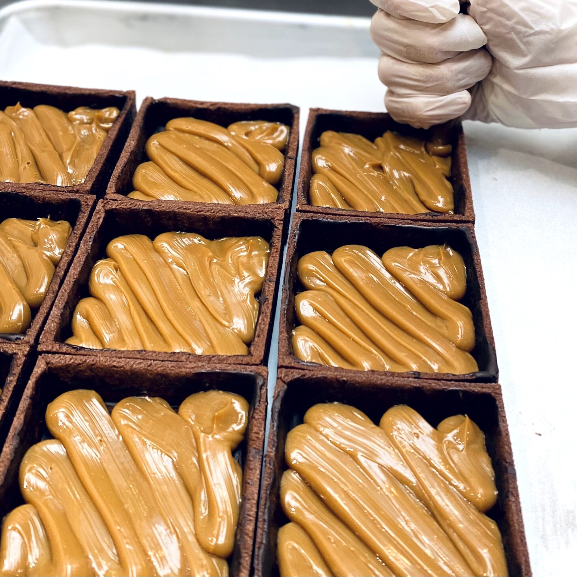 Close-up of caramel-filled chocolates in square boxes, with a gloved hand in the top right corner, on a white surface.
