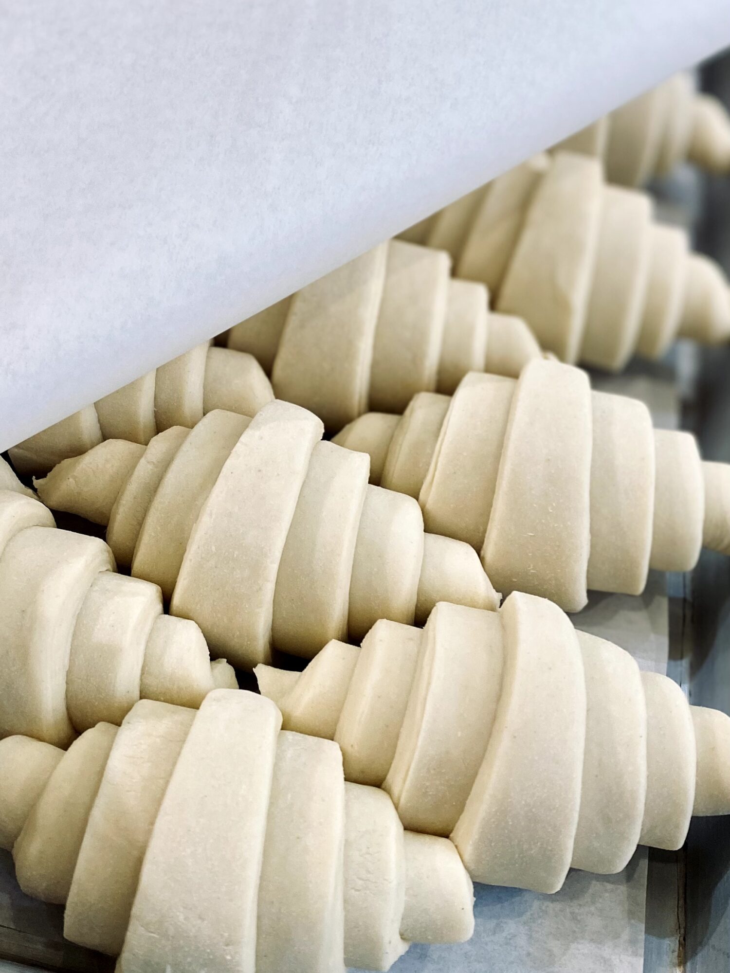 Rows of uncooked, spiral-shaped pastry dough on a baking sheet, partially covered by parchment paper.