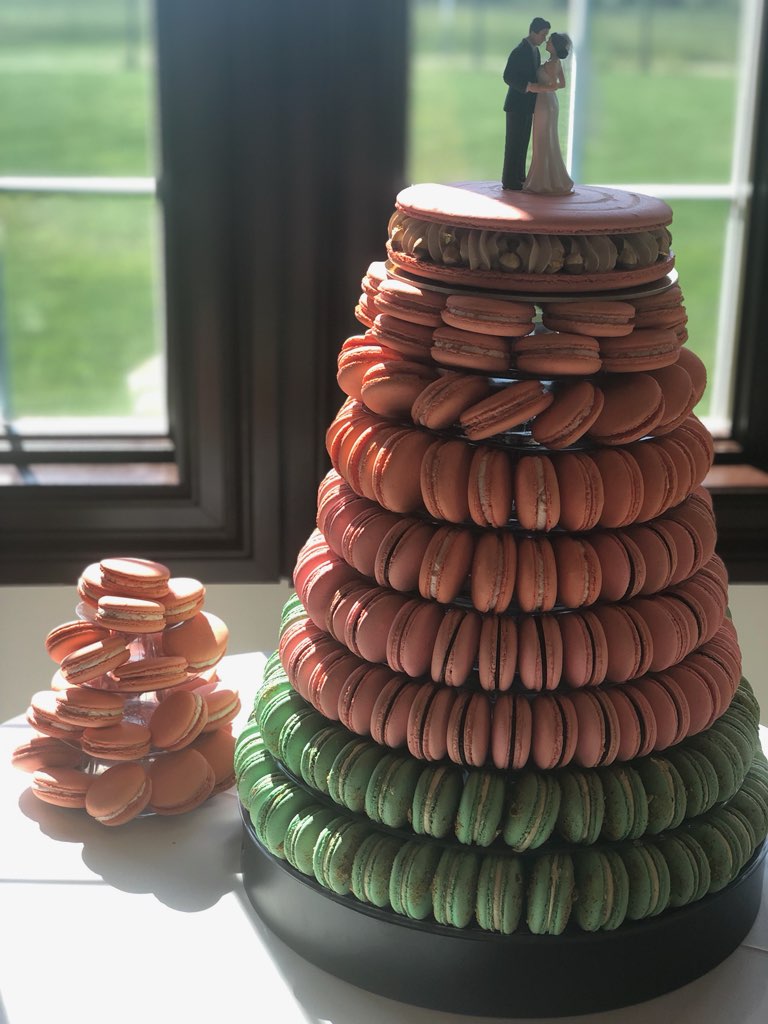 A tall stack of pink and green macarons with a wedding cake topper on top, placed on a black base near a window.