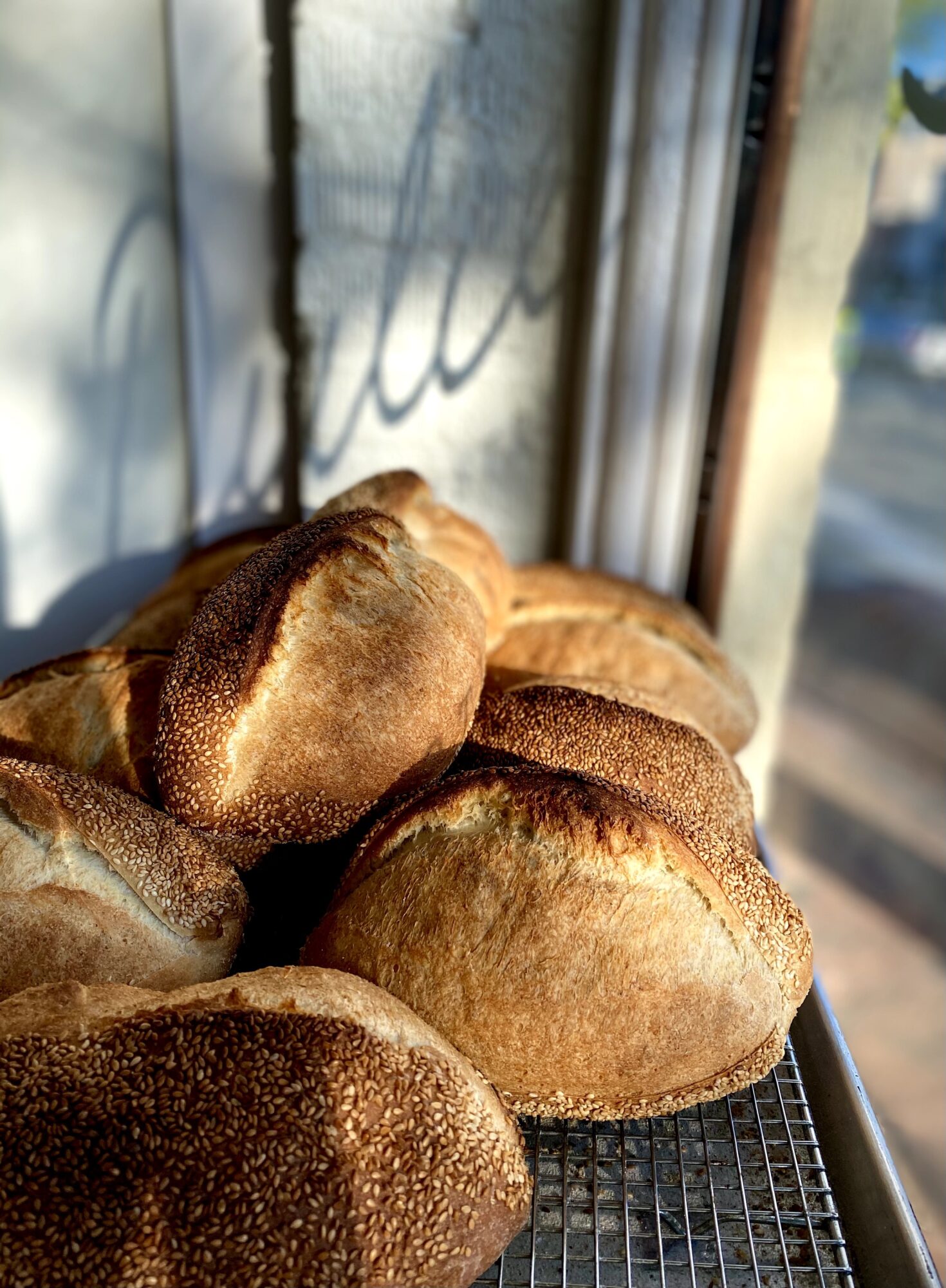 Several loaves of bread on a wire rack near a window with sunlight shining through, casting shadows on the wall.