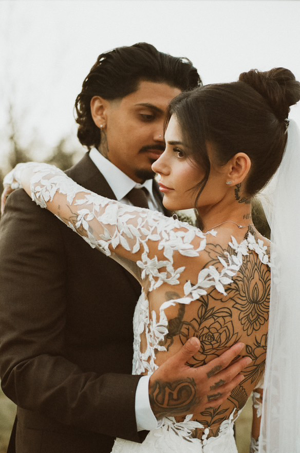 A couple in formal attire embrace closely, with the woman showing her back and intricate tattoo, and the man holding her.