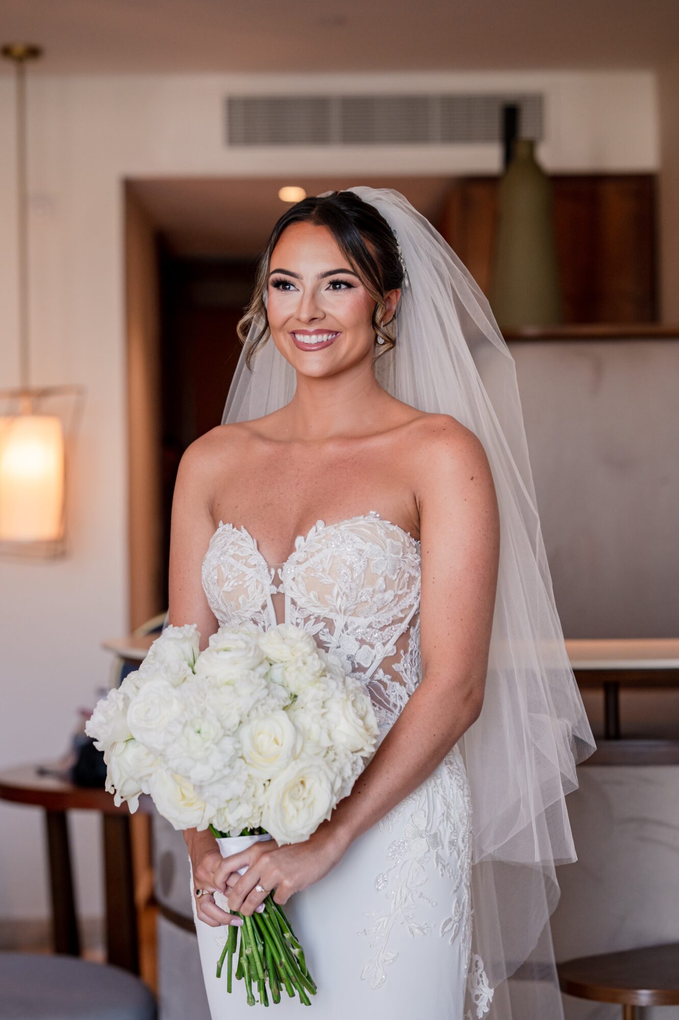 Bride in wedding dress holding bouquet, smiling indoors with veil and hair styled elegantly.