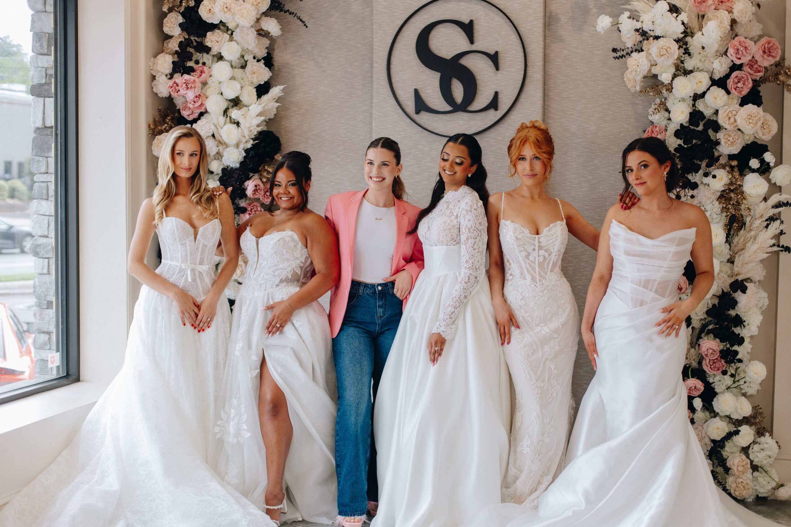 Six women in wedding dresses and casual attire stand in front of floral decorations and a logo, smiling.