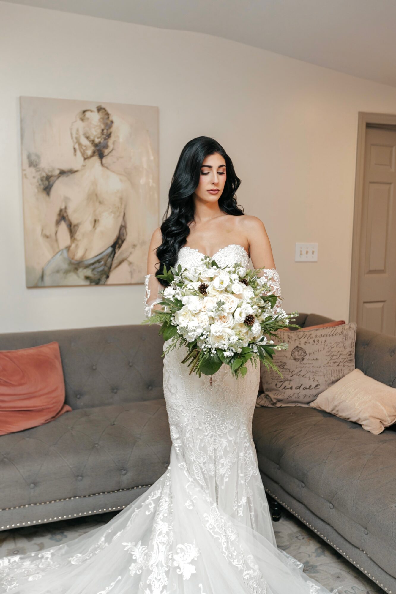Woman in wedding dress holding bouquet, standing in living room with artwork and couches.