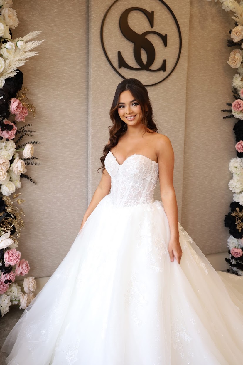 Young woman in a white wedding dress standing in front of floral decorations and a logo, smiling.