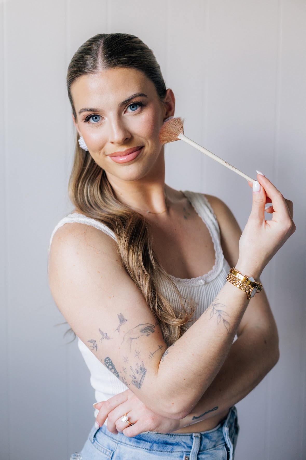 Young woman with long hair and tattoos, holding a makeup brush near her face, smiling, against a light background.