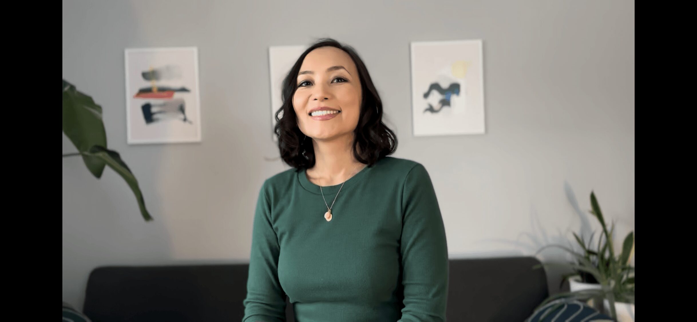 Woman with dark hair smiling, wearing a green top, sitting in a room with artwork on the wall.