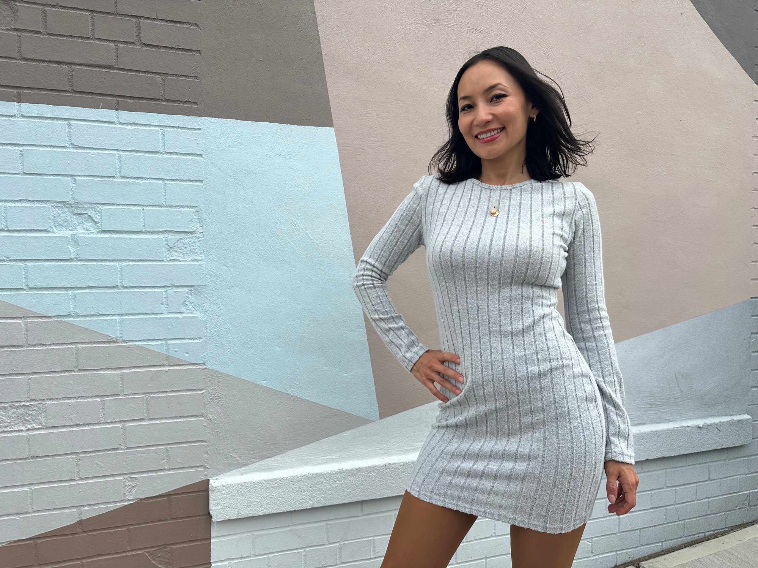 Woman in a light gray, long-sleeved, ribbed dress standing outdoors against a geometric mural background, smiling with one hand on her hip.