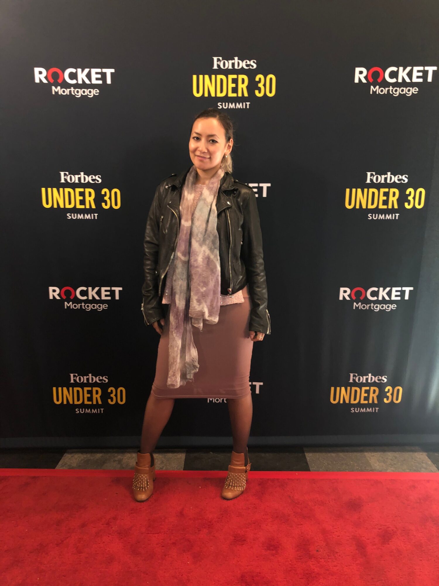 Woman standing on red carpet in front of black backdrop with text, wearing a light-colored dress and a dark jacket.