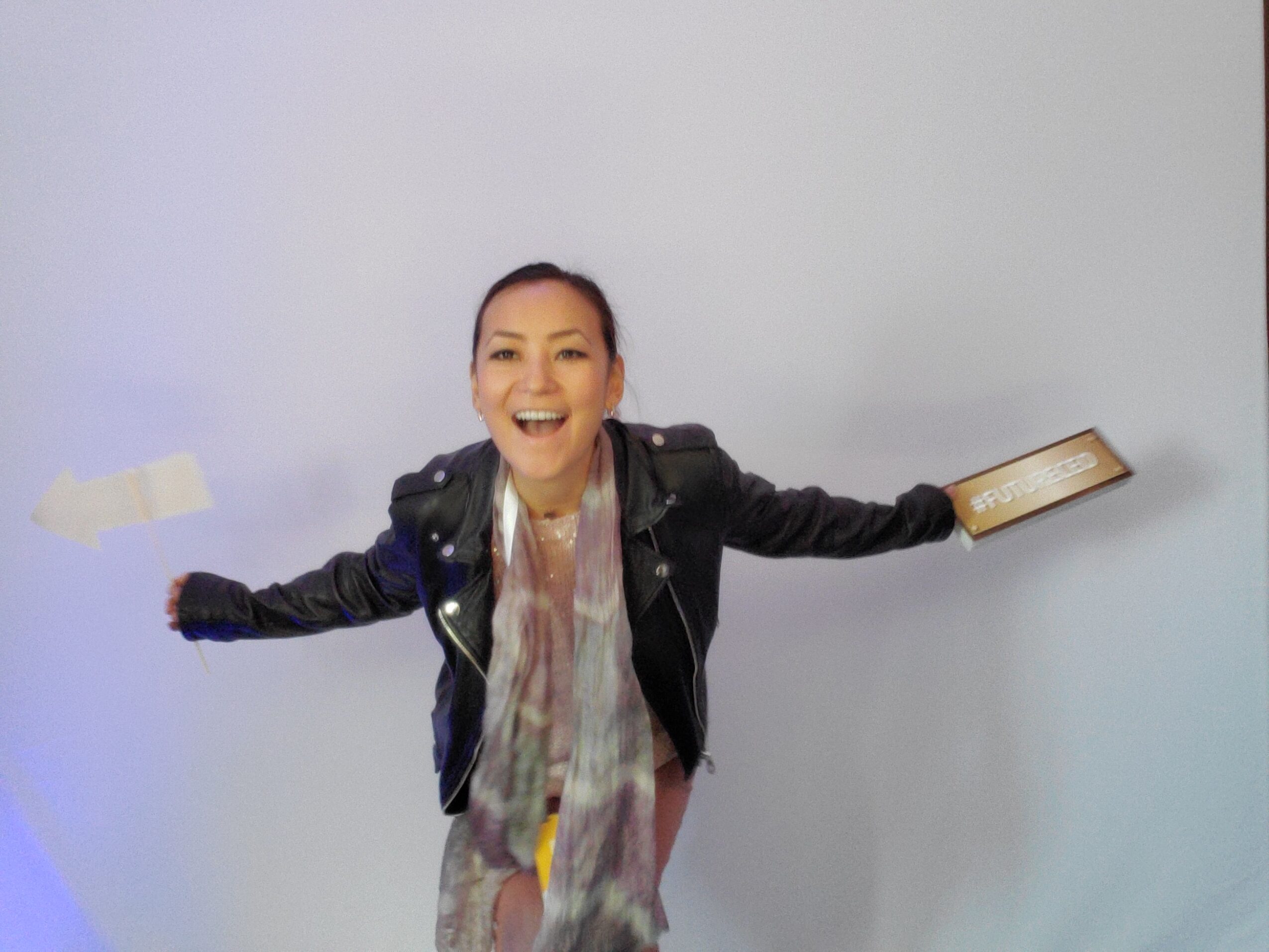 Person with arms outstretched, smiling, holding a book in one hand, against a plain background.
