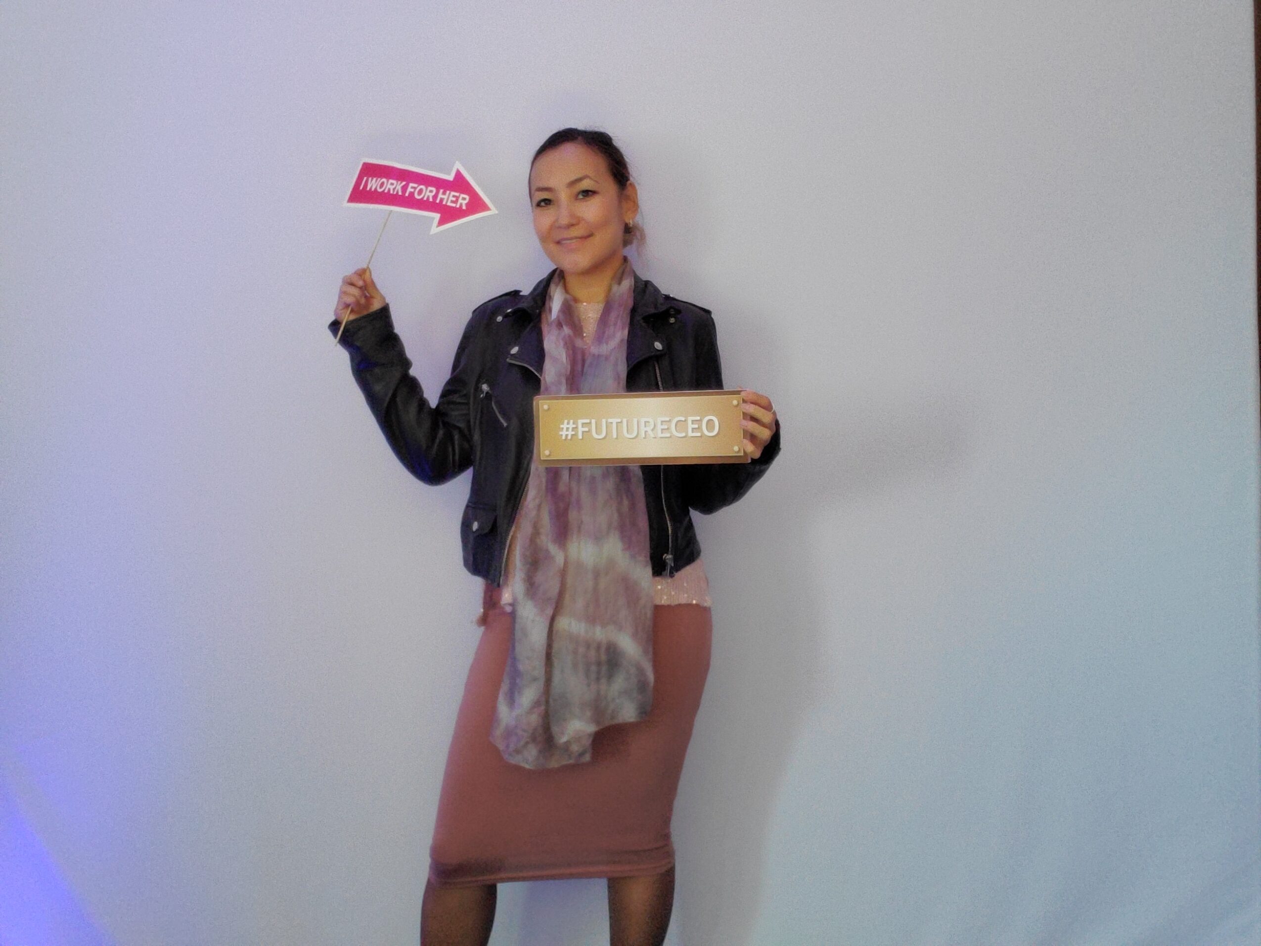 Woman holding a sign and a cardboard with text, standing against a plain wall.
