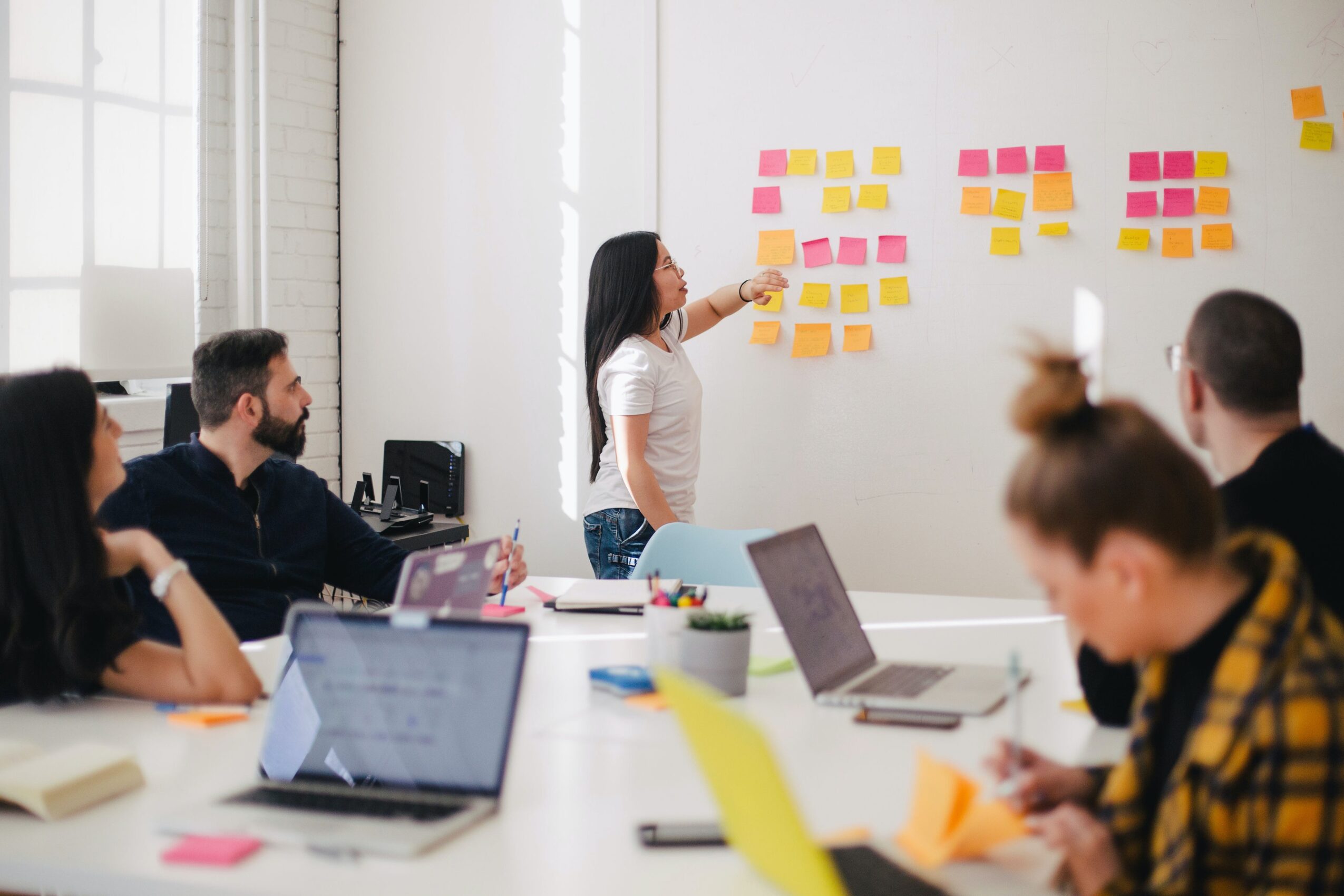 Group of people in a meeting room with a woman presenting sticky notes on a wall, laptops and notebooks on table