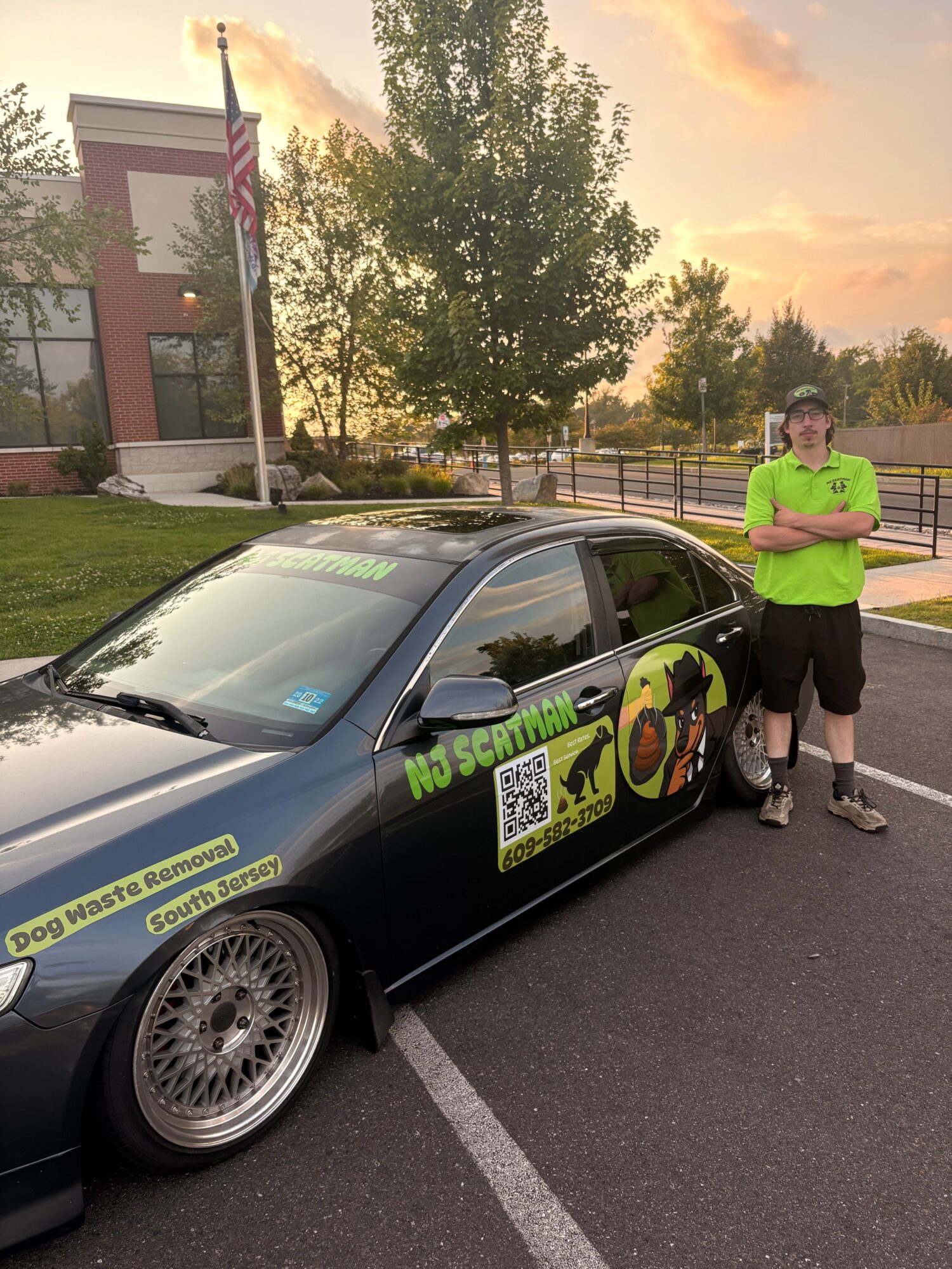 Person in bright green shirt and dark pants standing next to a black car with colorful decals, in a parking lot at sunset.