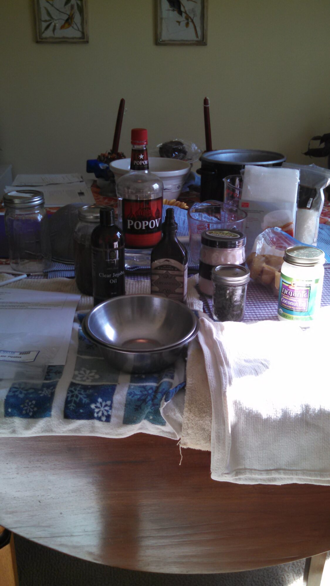 Table with jars, bottles, and containers, covered with a cloth, in a room with framed pictures on the wall.