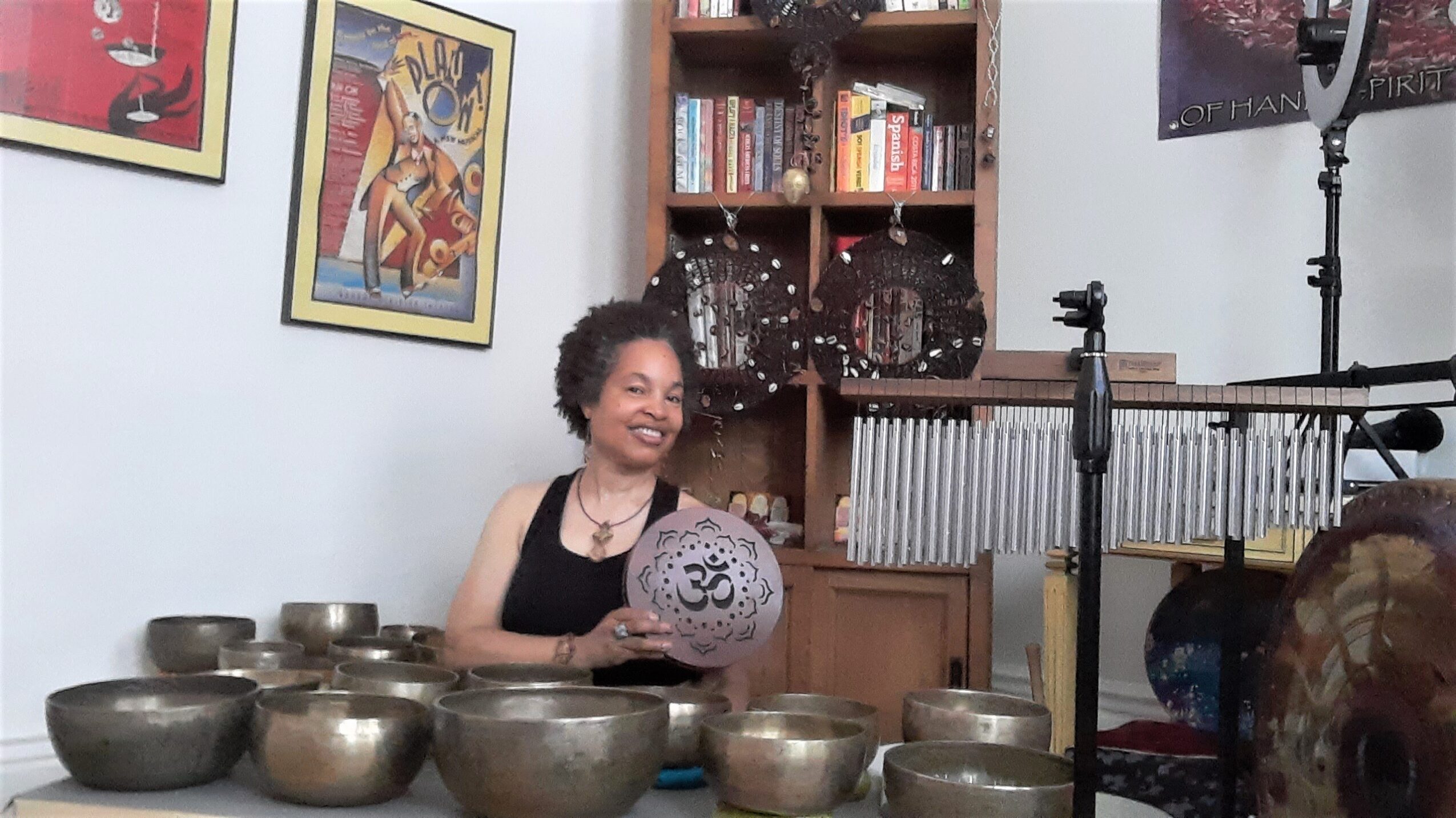 Woman smiling, holding a decorative object, surrounded by singing bowls, in a room with artwork and musical instruments.