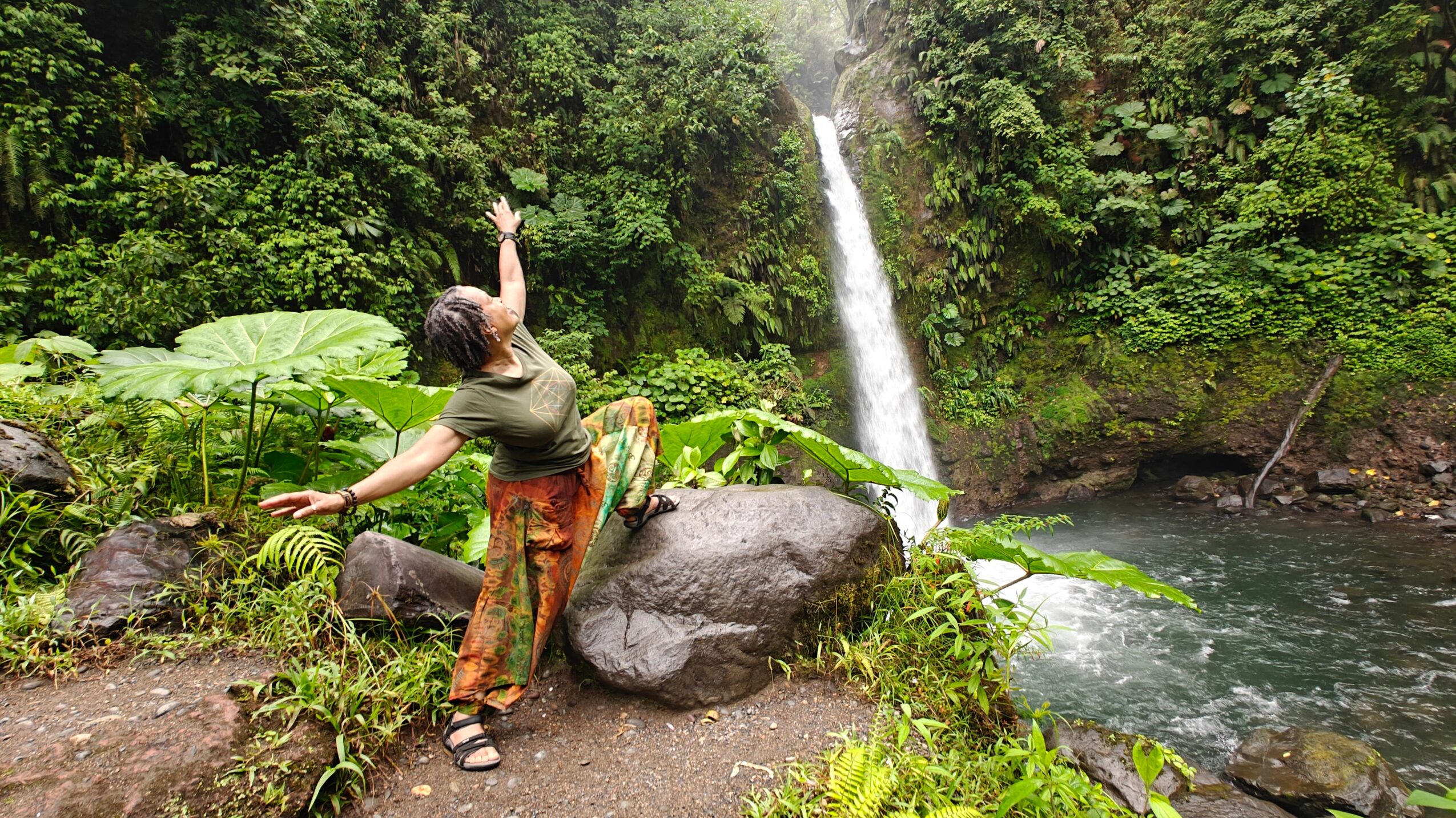 Person sitting on a large rock near a waterfall in a lush green forest, reaching upward with one arm.