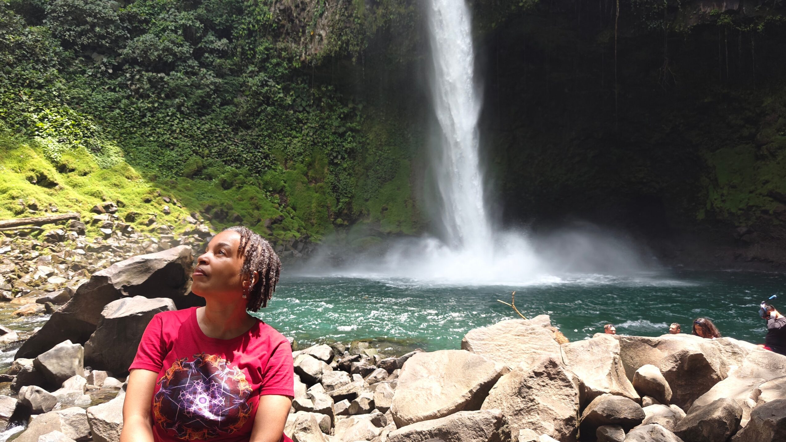 Person in red shirt sitting on rocks near waterfall and river, surrounded by greenery