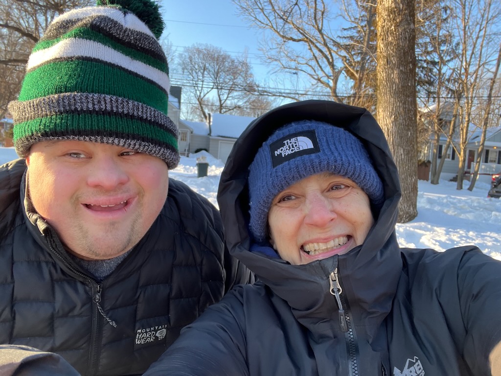 Two people outdoors in winter, smiling, wearing winter hats and jackets, with snow and trees in the background.