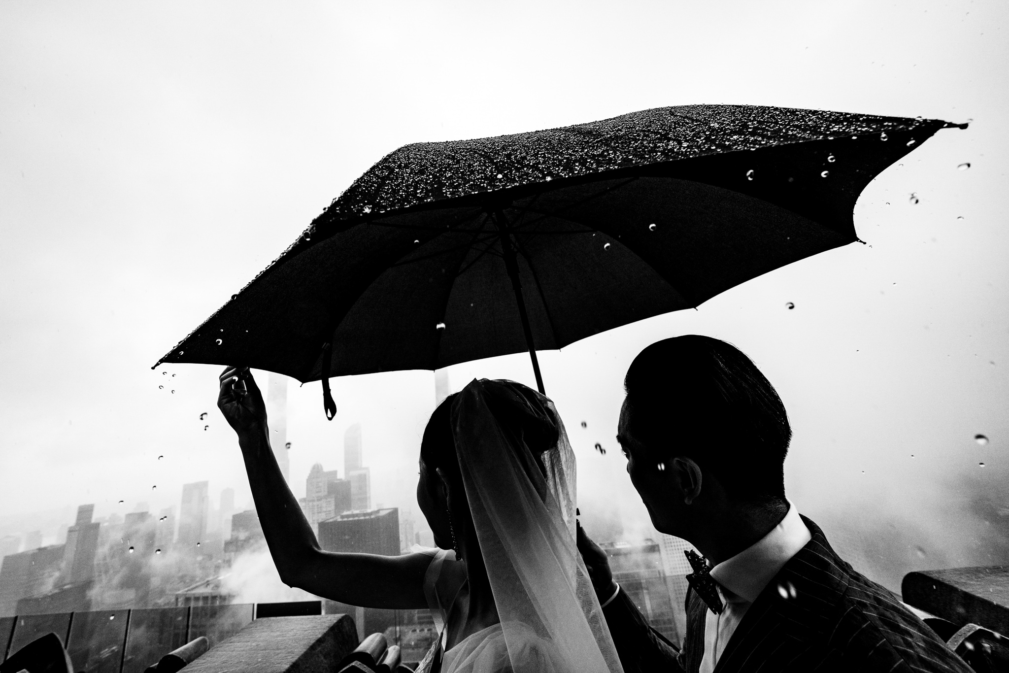 Two people under an umbrella on a rainy day with a city skyline in the background, black and white.