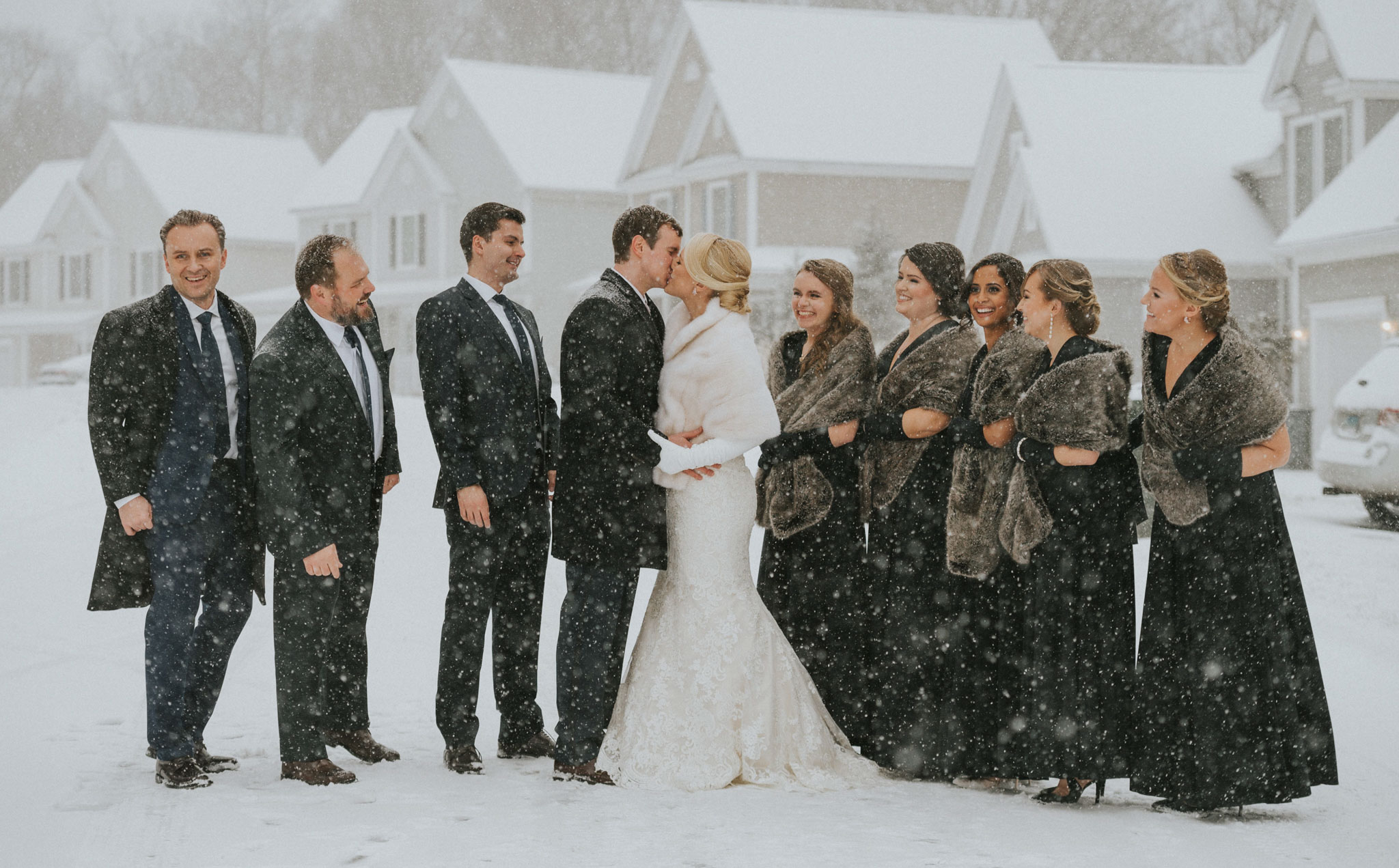 A wedding party stands outdoors in snow, with a bride and groom in the center, surrounded by friends and family.