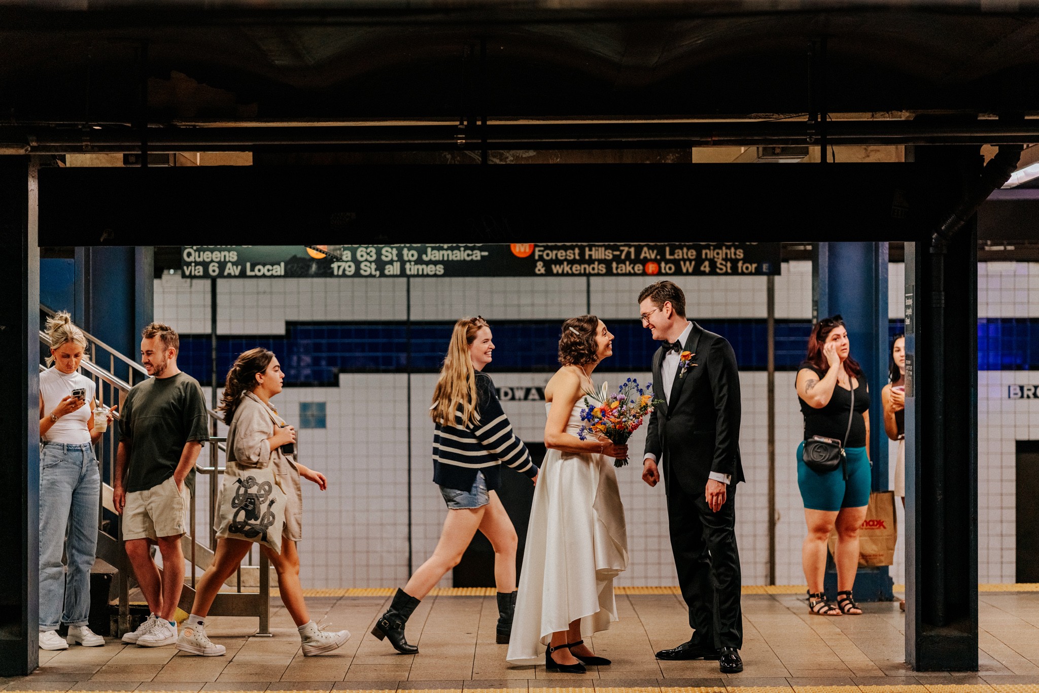 People standing and walking on a subway platform, with a couple in formal attire in the center, some waiting or talking.