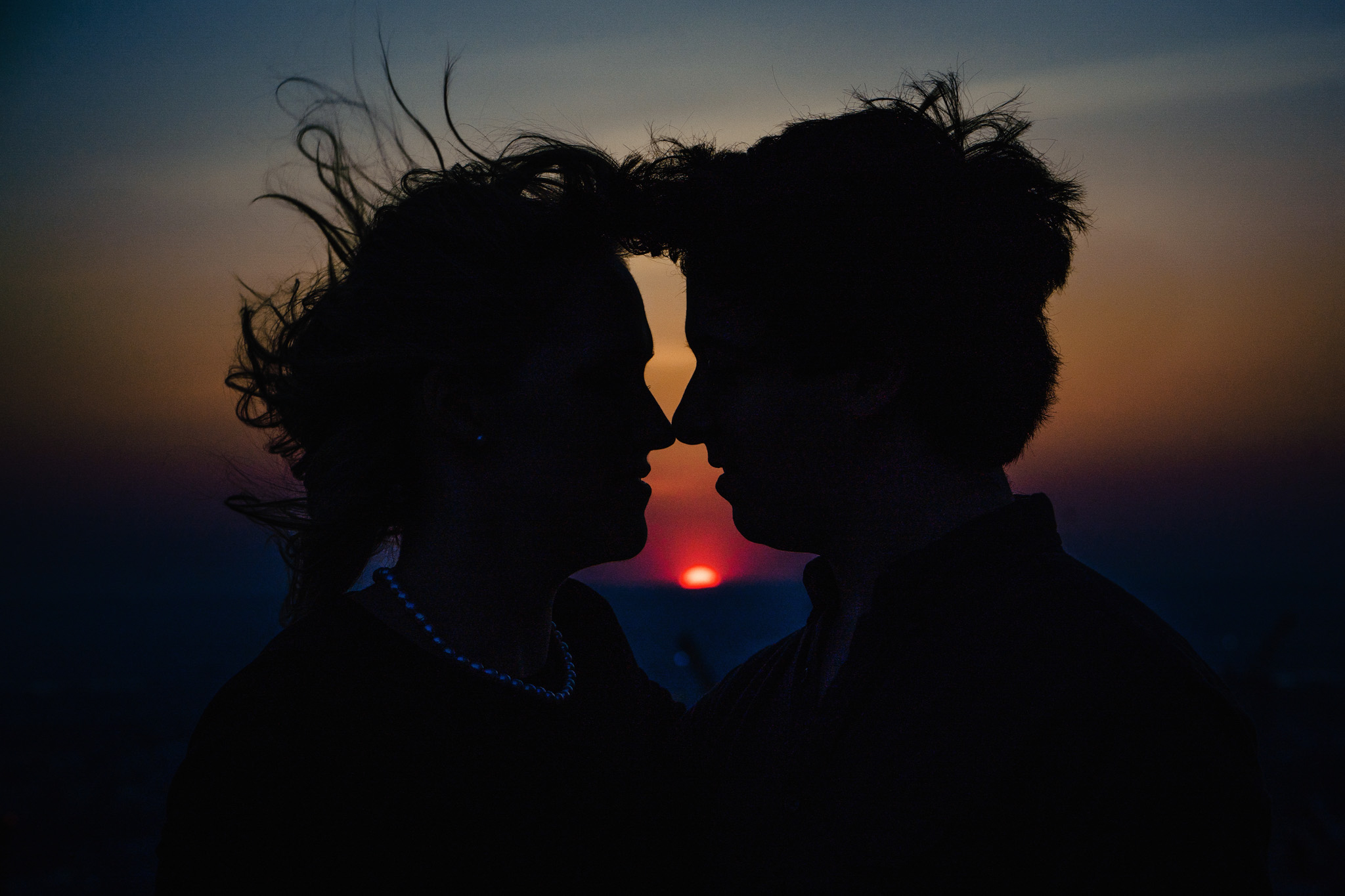 Two people face each other at sunset with their silhouettes visible, and the sun setting behind them.