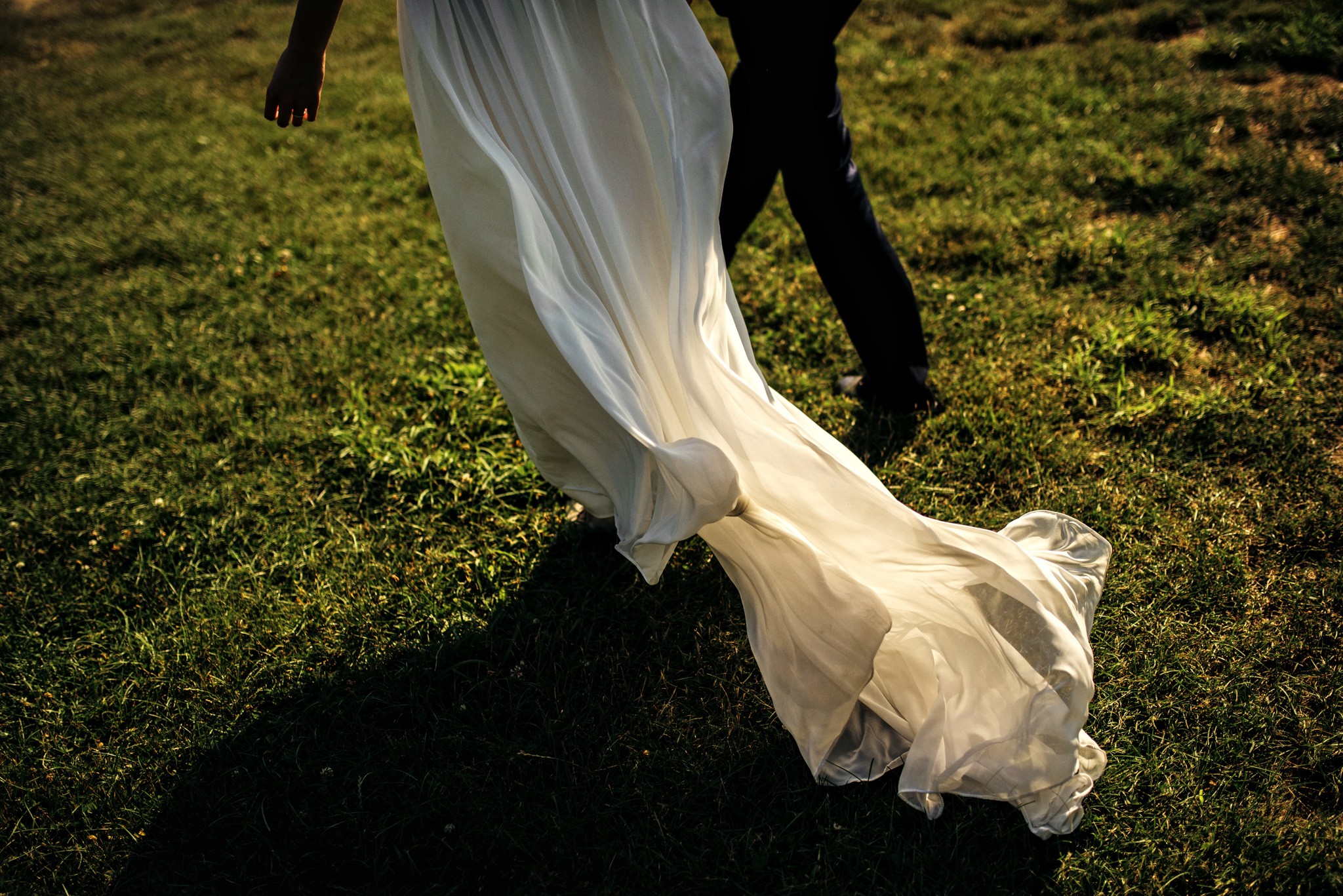 Person in a flowing white dress walking on grass, with another person nearby, sunlight casting shadows.