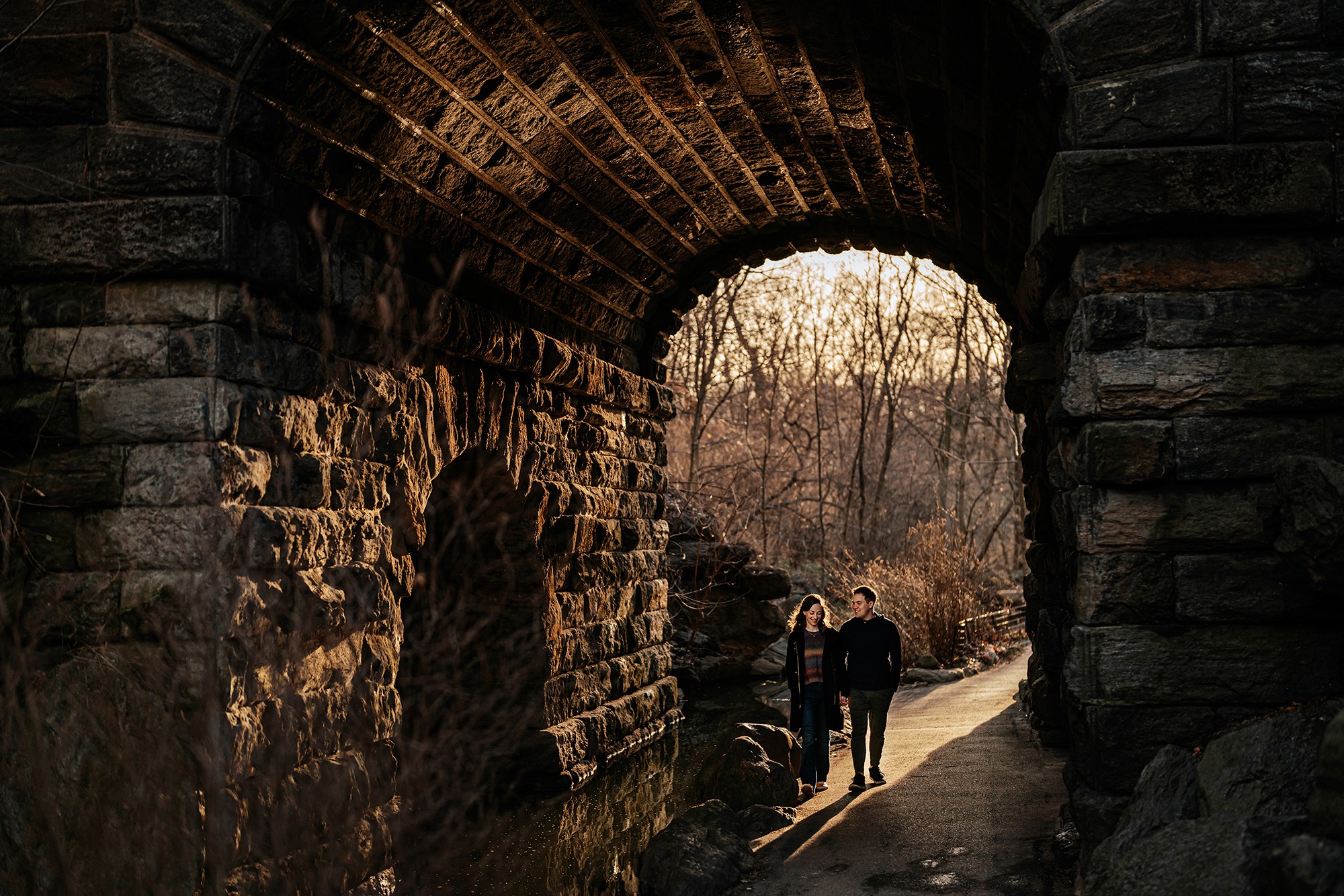 Three people walk through a stone tunnel with an opening showing trees and sky outside.