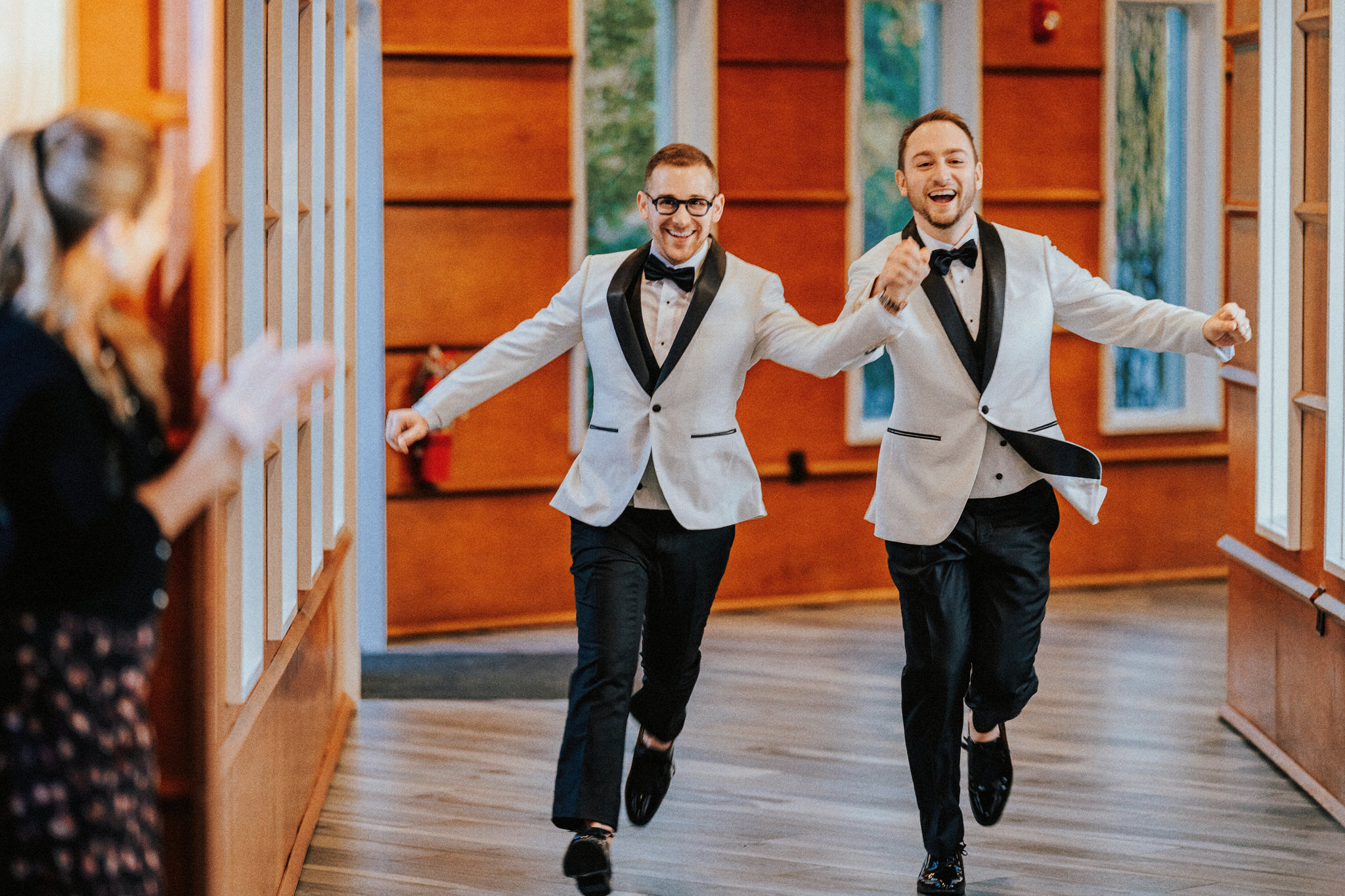 Two men in tuxedos run through a hallway, smiling and holding hands, with a woman clapping nearby.