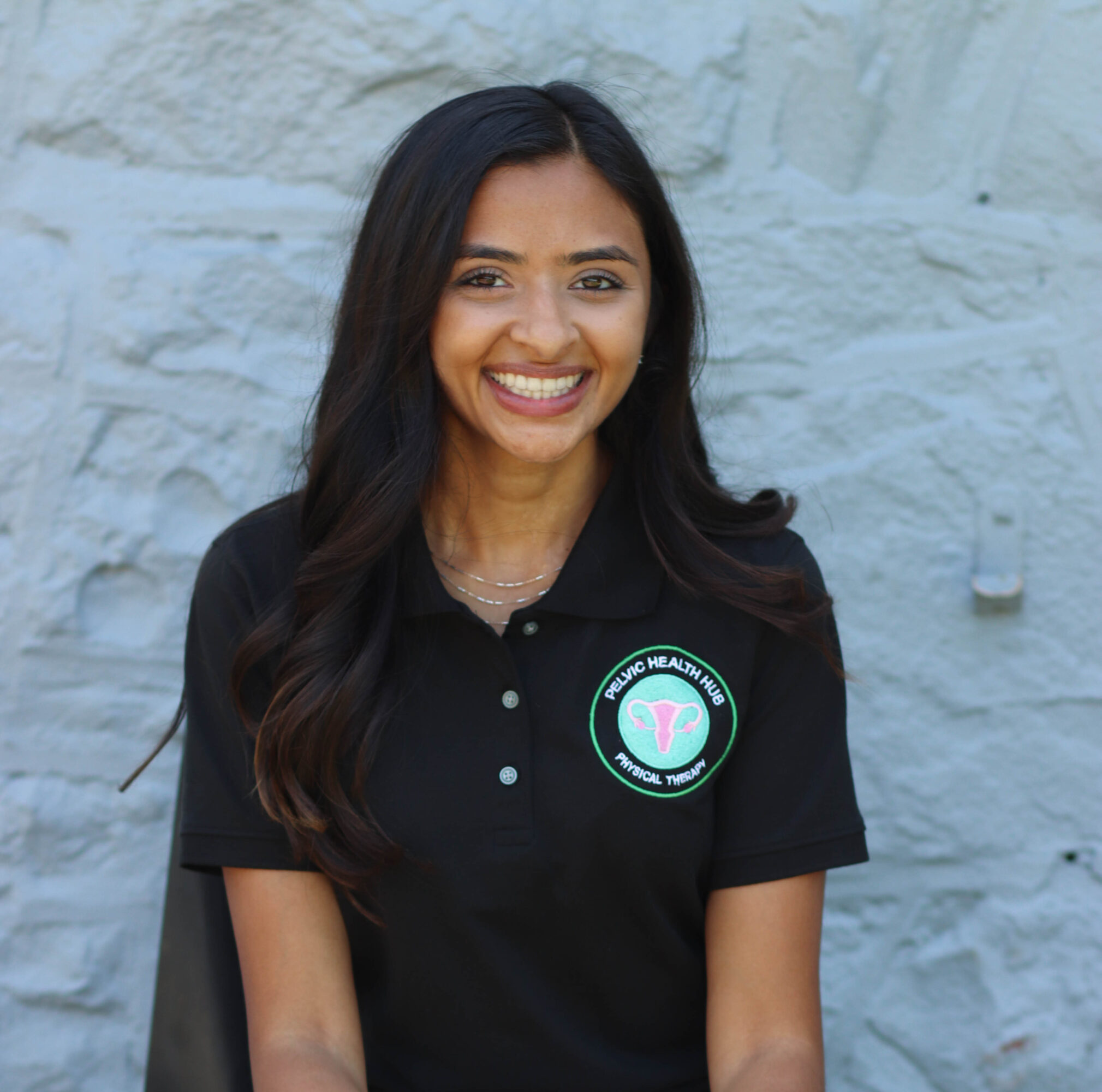 Smiling woman with long dark hair wearing a black polo shirt with a logo, standing against a light-colored stone wall.